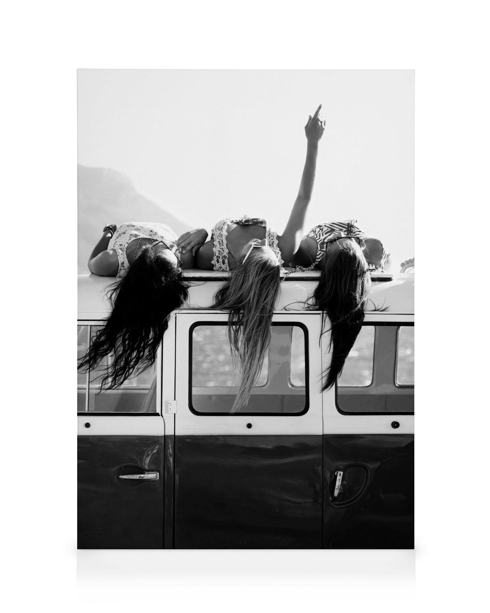 Black and white canvas print of three women lying on a car roof with hair flowing, one arm raised.