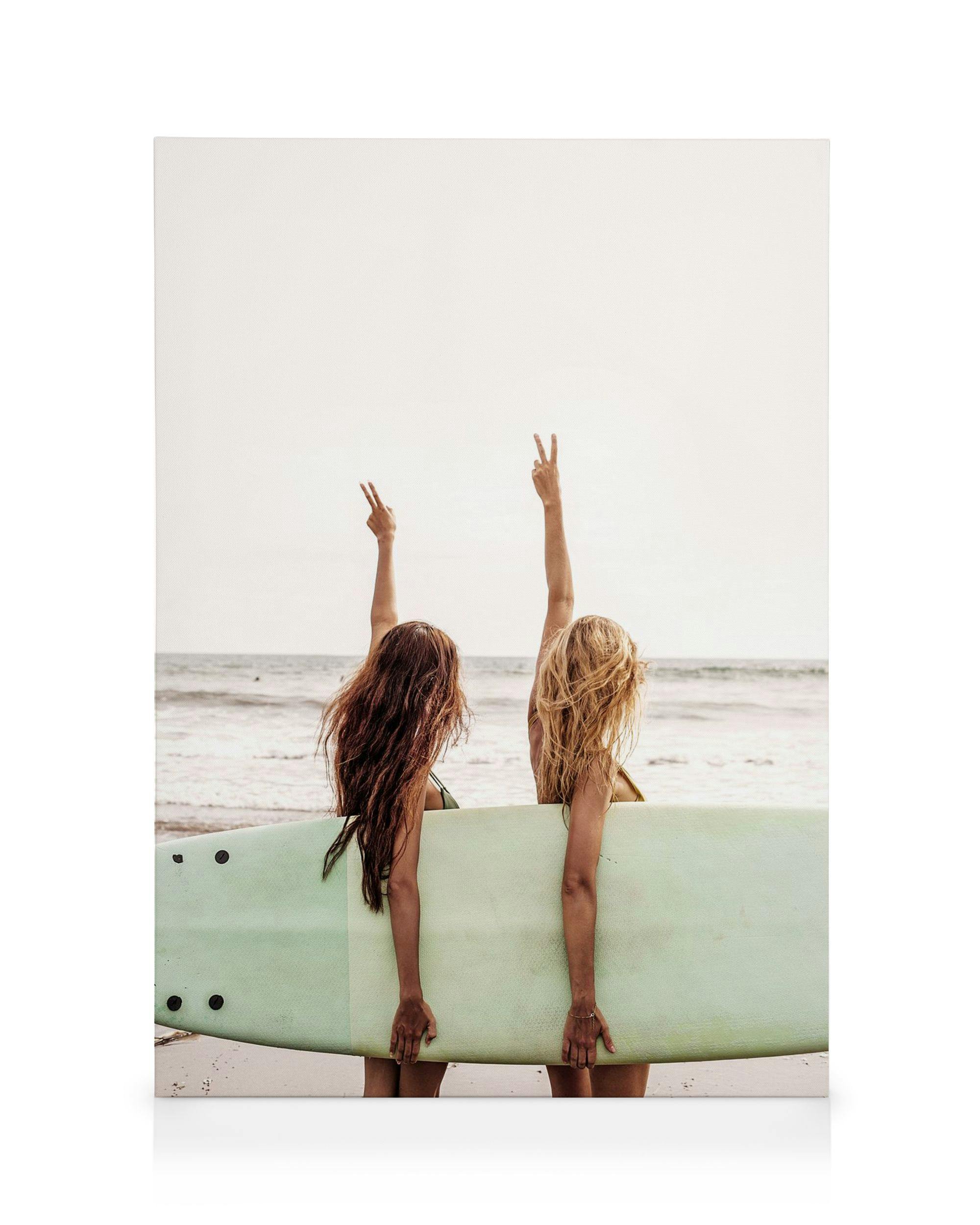 Two surfer girls with long hair, seen from behind, holding a surfboard on the beach and making peace signs towards the ocean.