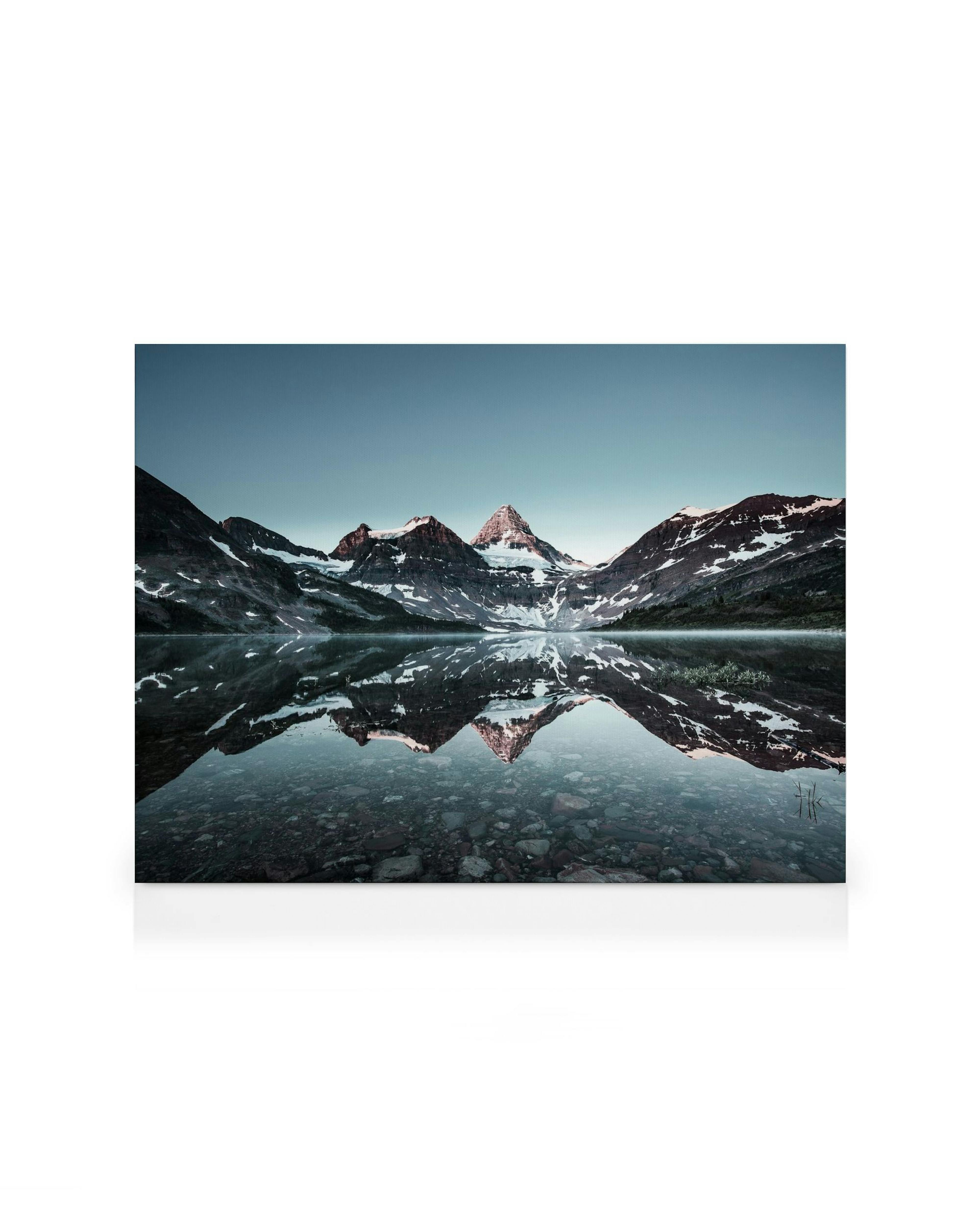 Bergmeer canvas: besneeuwde bergen weerspiegeld in water onder lichtblauwe lucht, rotsen zichtbaar op de bodem.