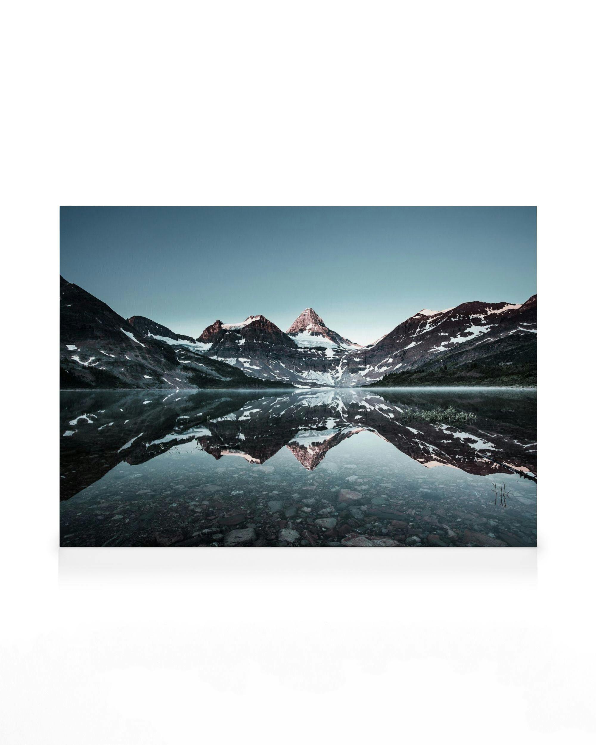 Bergmeer canvas: besneeuwde bergen weerspiegeld in water onder lichtblauwe lucht, rotsen zichtbaar op de bodem.