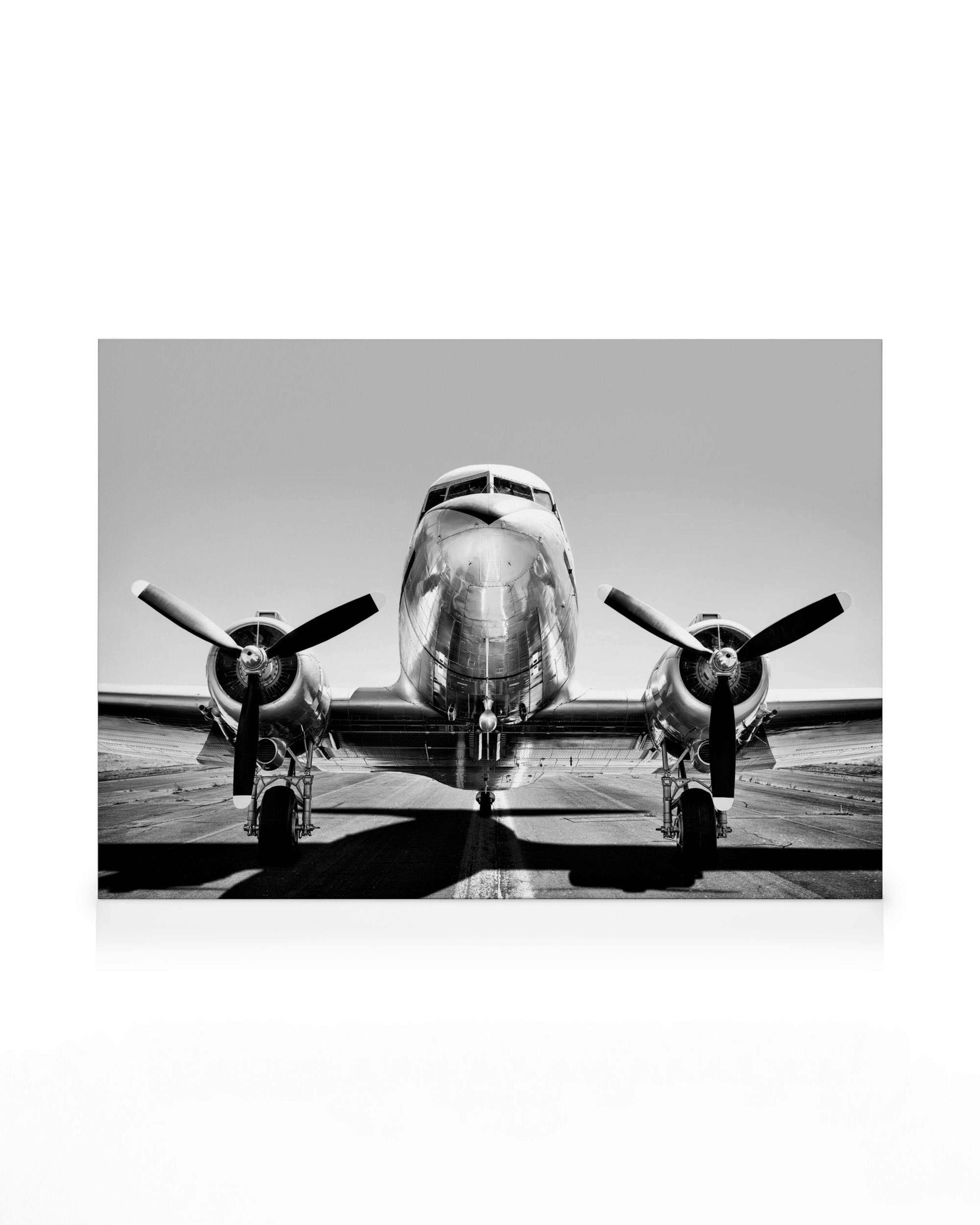 Zwart-wit canvas van een vintage vliegtuig op een landingsbaan, met glanzende propellers en romp tegen een heldere lucht.