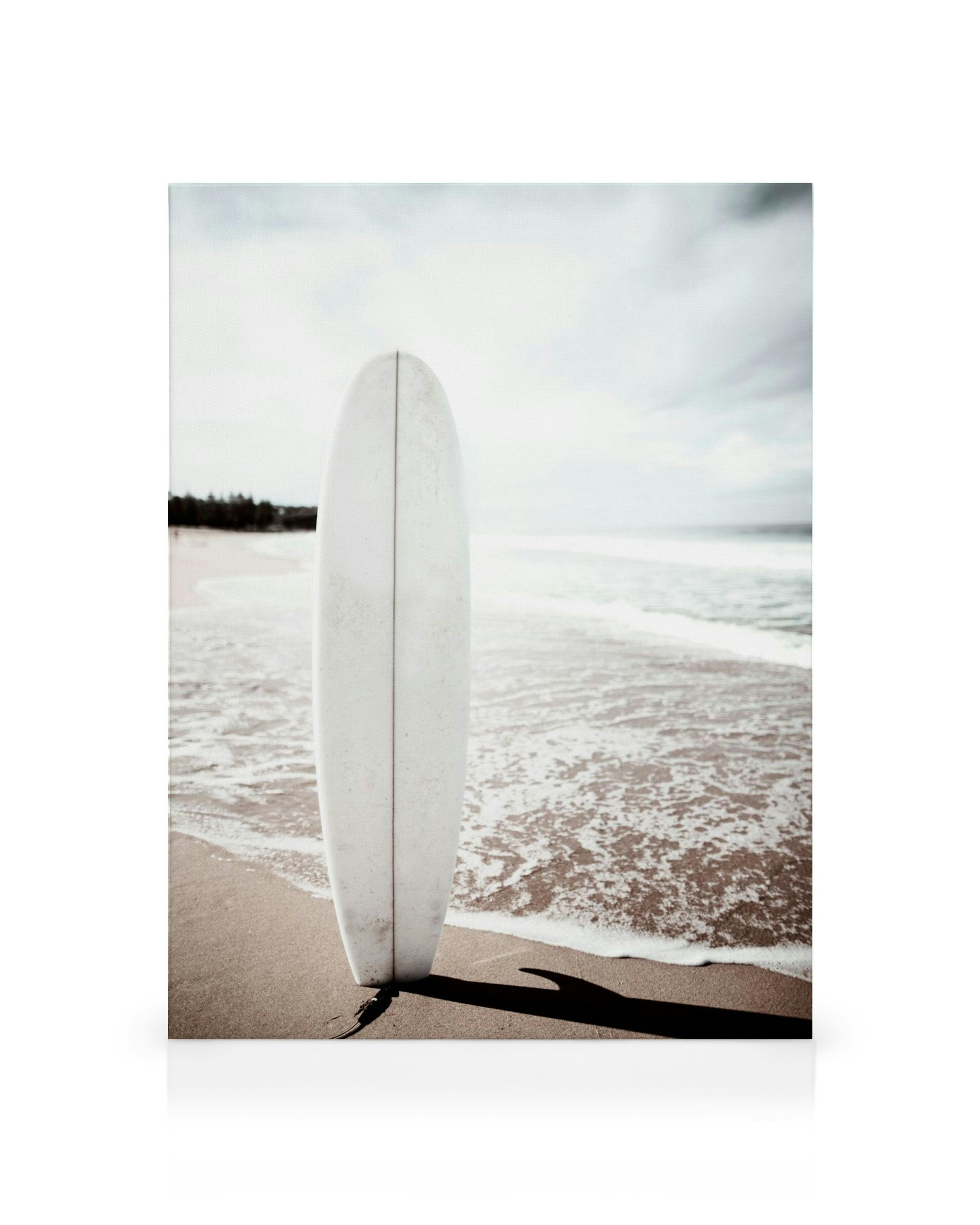 White surfboard on sandy beach with sea foam and shadow, coastal wall art print.