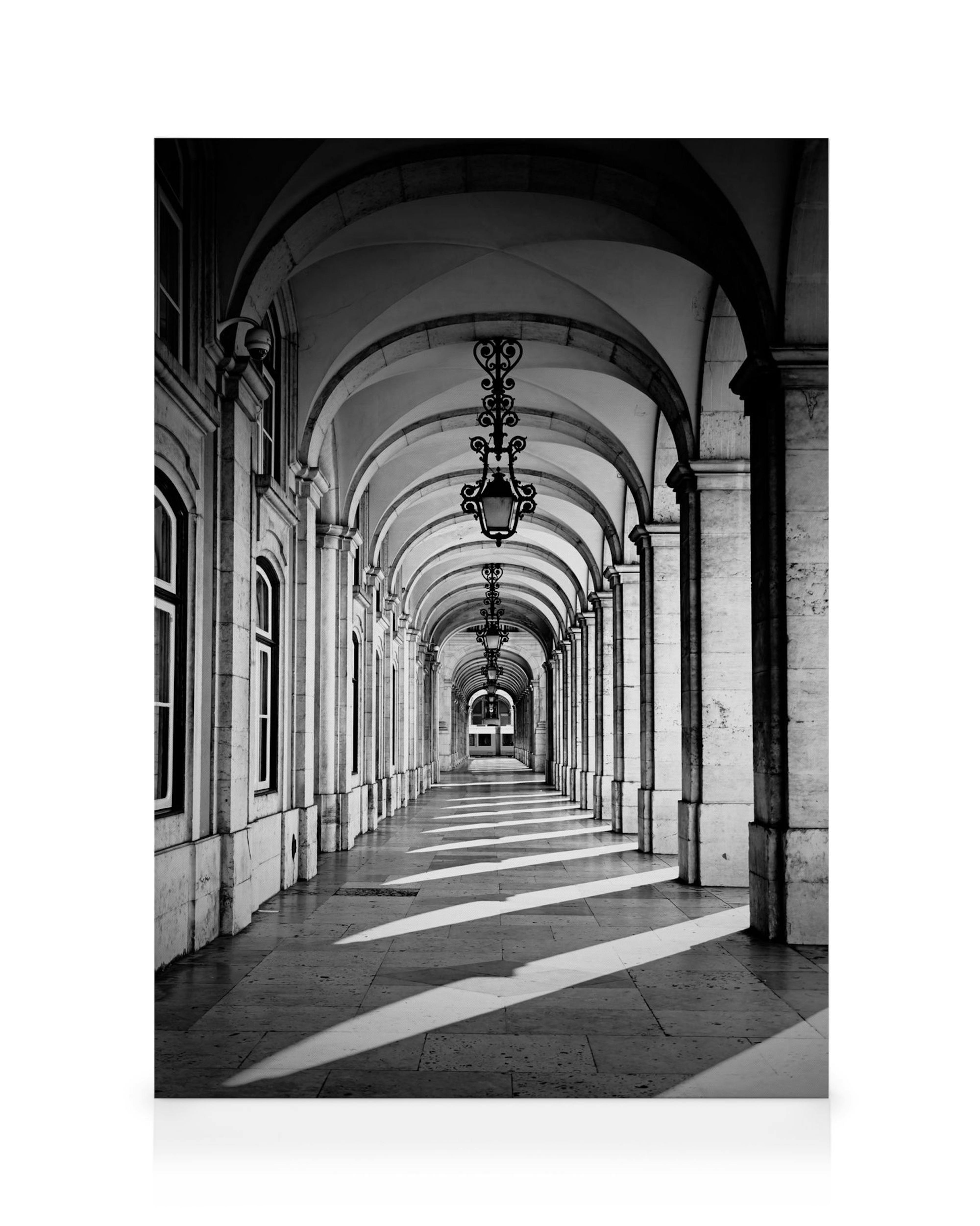 Black and white photograph of a long arched corridor with ornate hanging lamps, sunlight casting dramatic shadows on the floor.