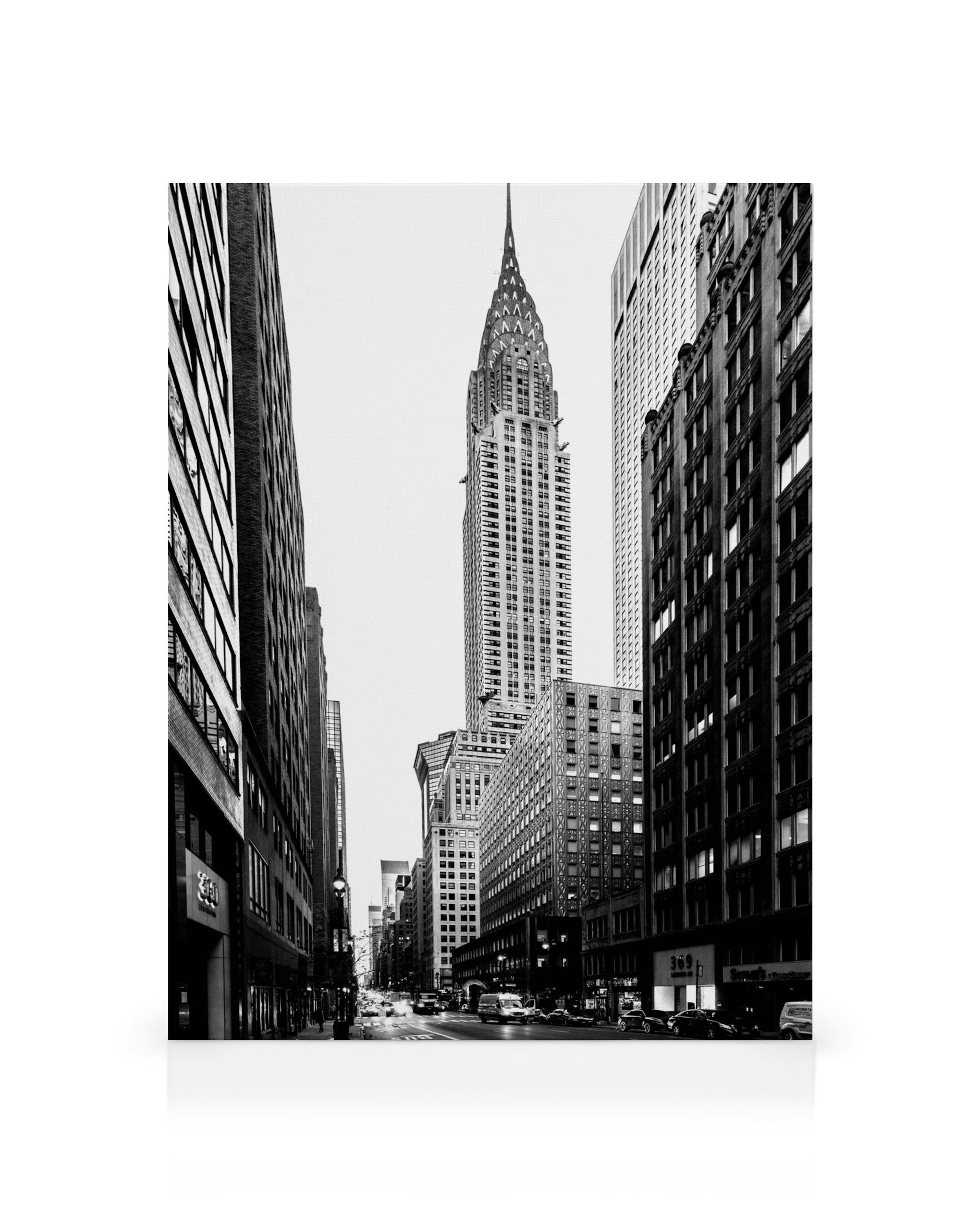 Mustavalkoinen katukuva New Yorkista, Chrysler Building hallitsee horisonttia pilvenpiirtäjien ja vilkkaan kadun keskellä.