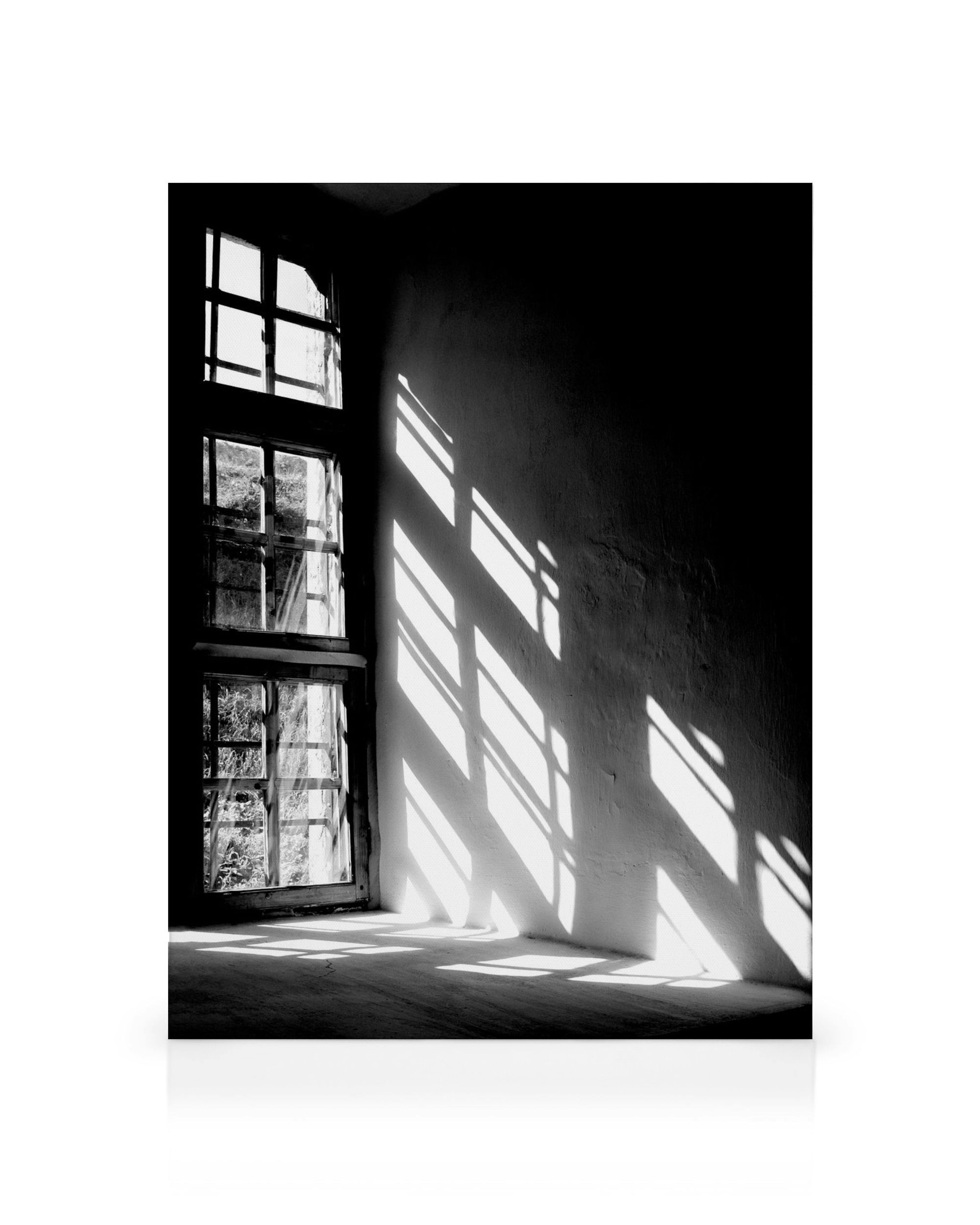 Schwarz-Weiss-Fotografie: Sonnenlicht fällt durch ein Fenstergitter und wirft Schatten auf eine Wand und den Boden.