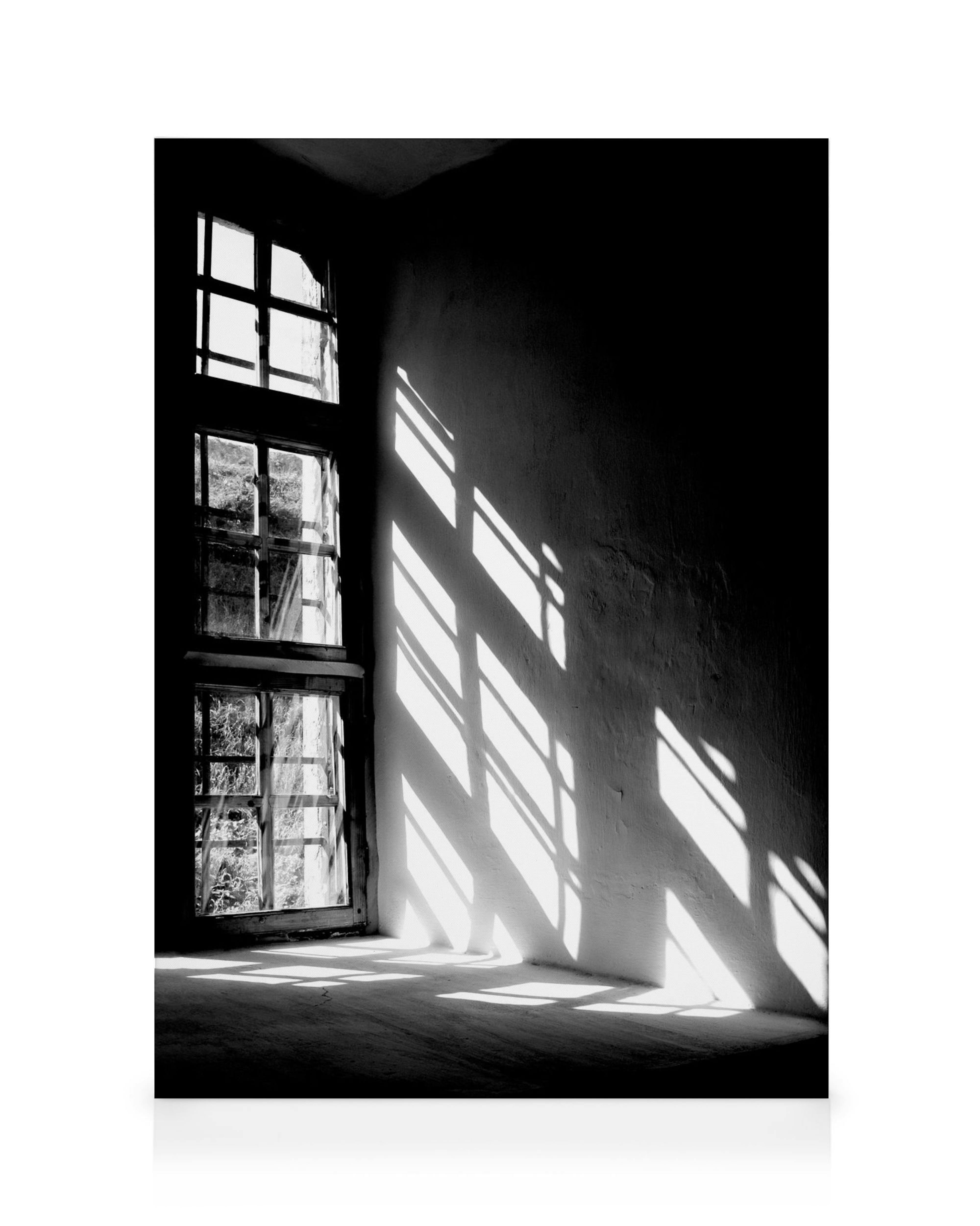Black and white image of a window with light streaming through, casting dramatic shadows on the wall and floor. Sunlight creates