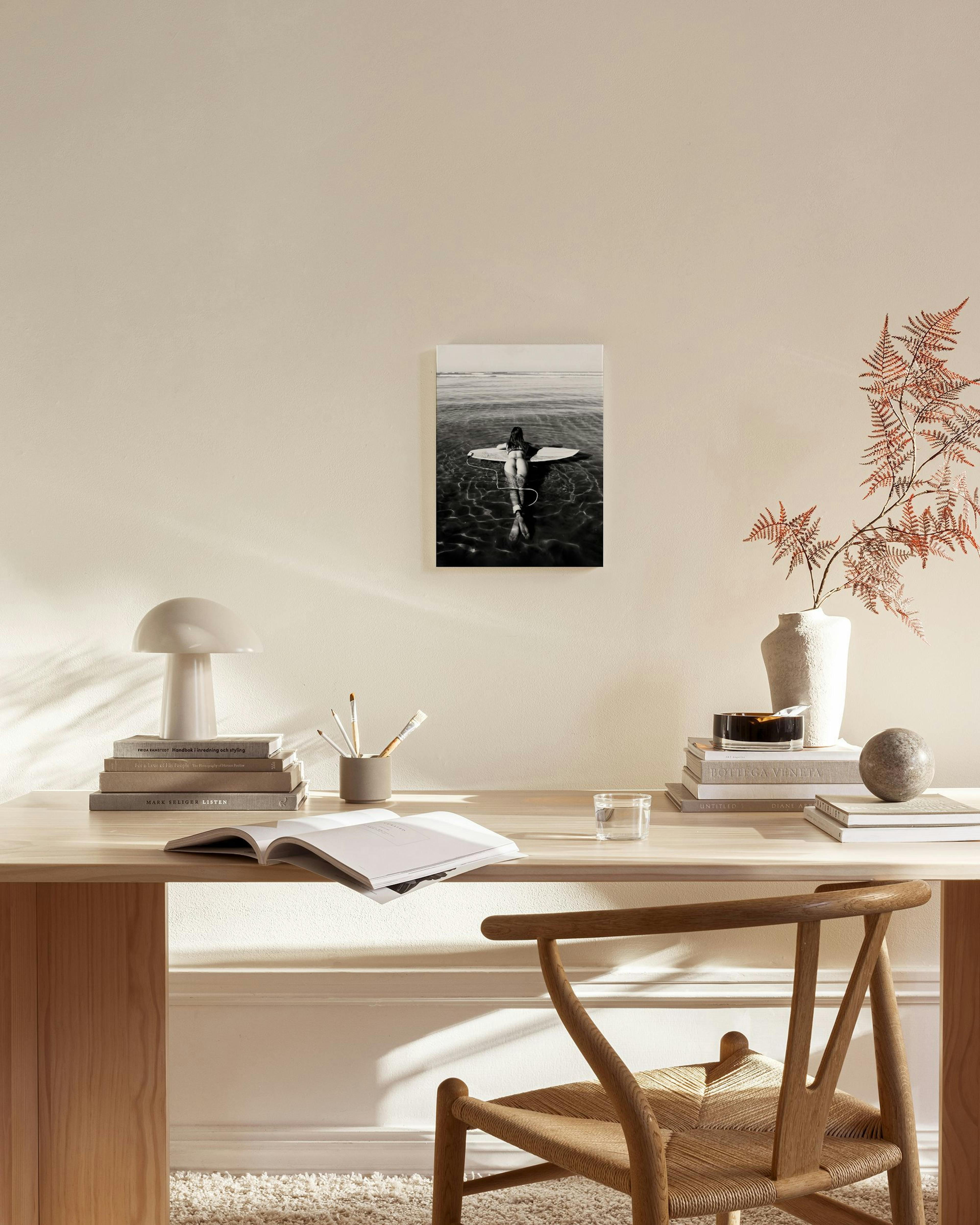 A black and white canvas print of a person on a surfboard in calm water, displayed in a bright living room with a light wood cha