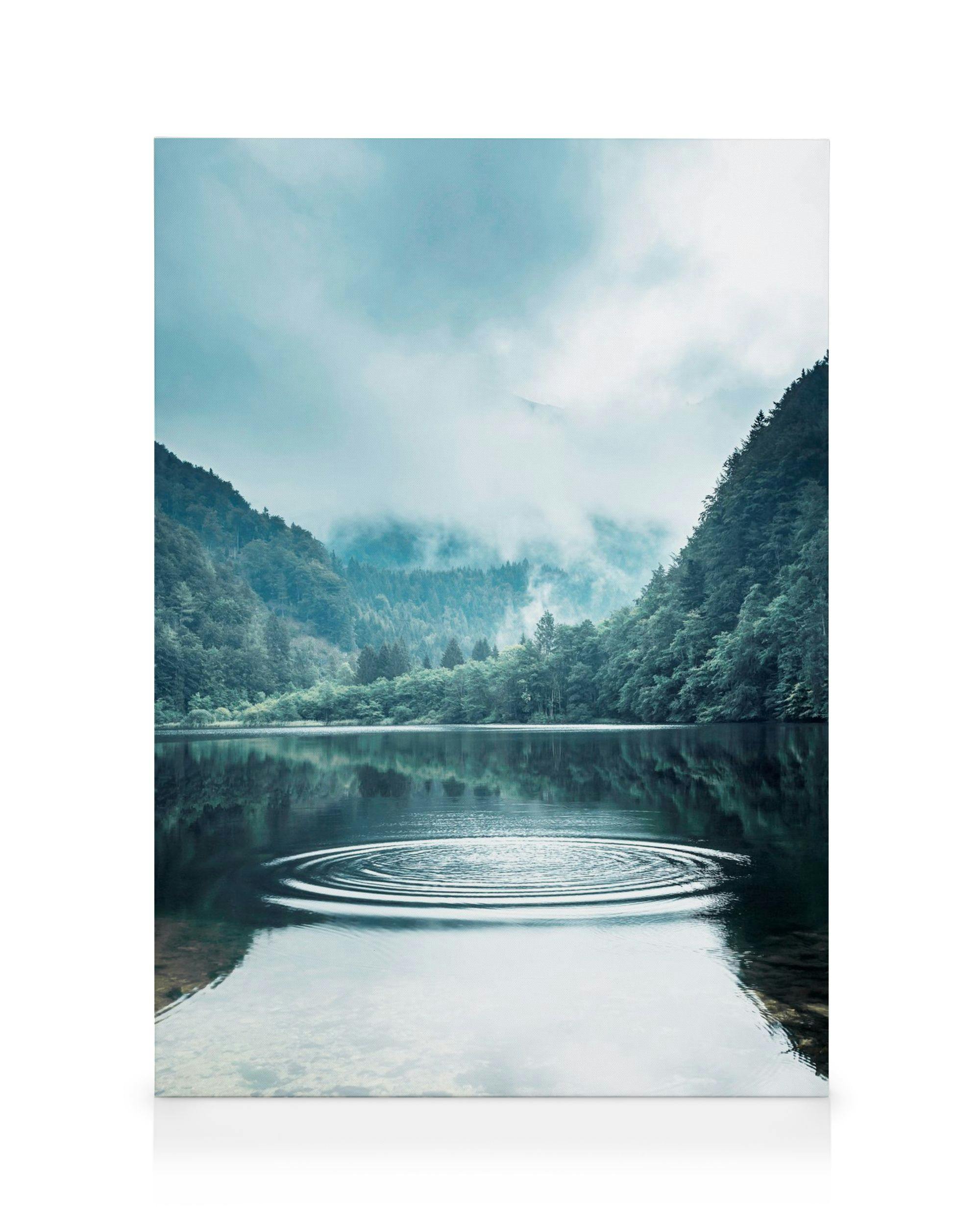 Photograph of a serene lake with ripple rings on the water, surrounded by a misty forest and mountains under a cloudy sky.