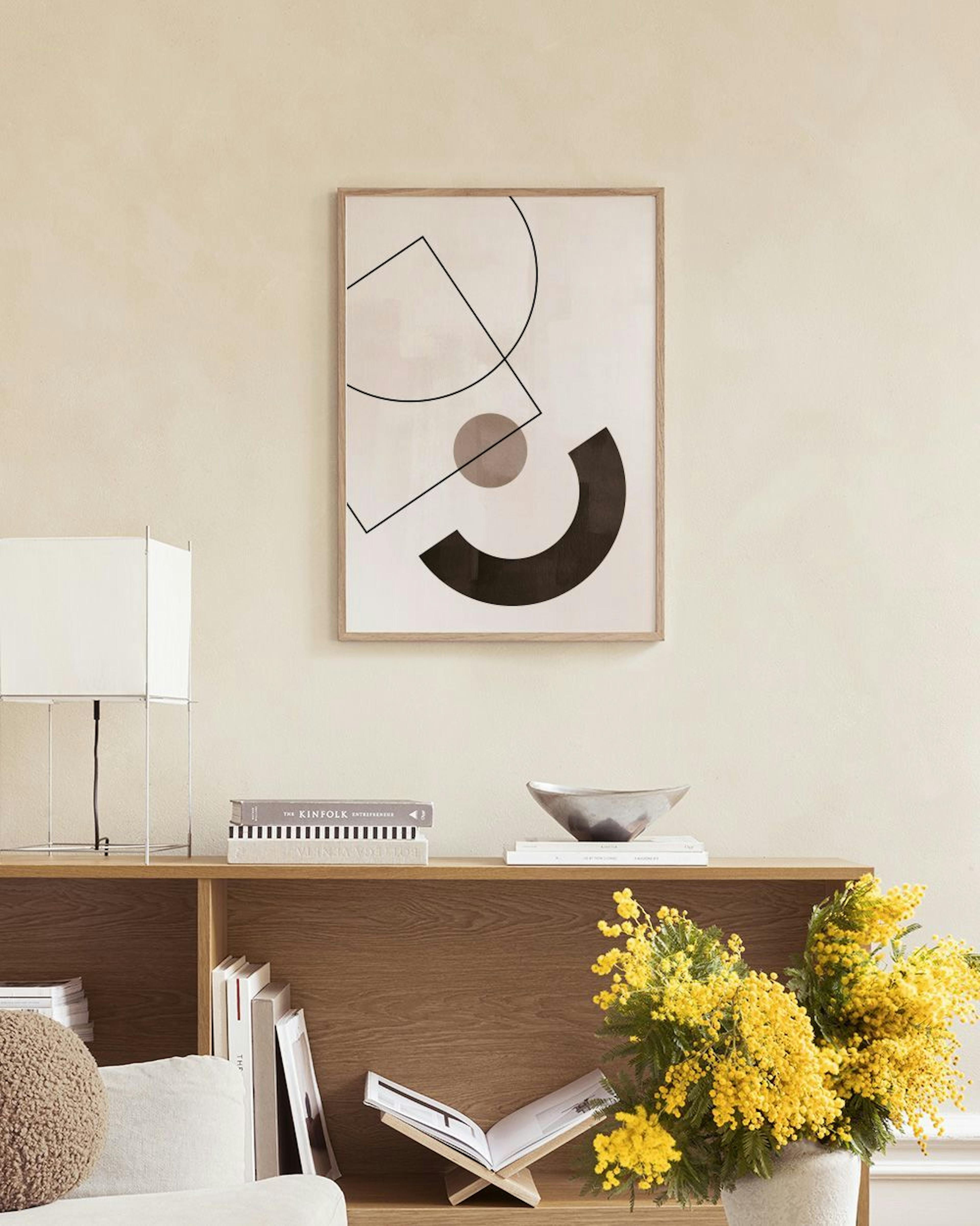 Poster mit geometrischen Formen in Schwarz und Grau, hängt über einem beigen Sessel, neben einer Vase mit Pampasgras.