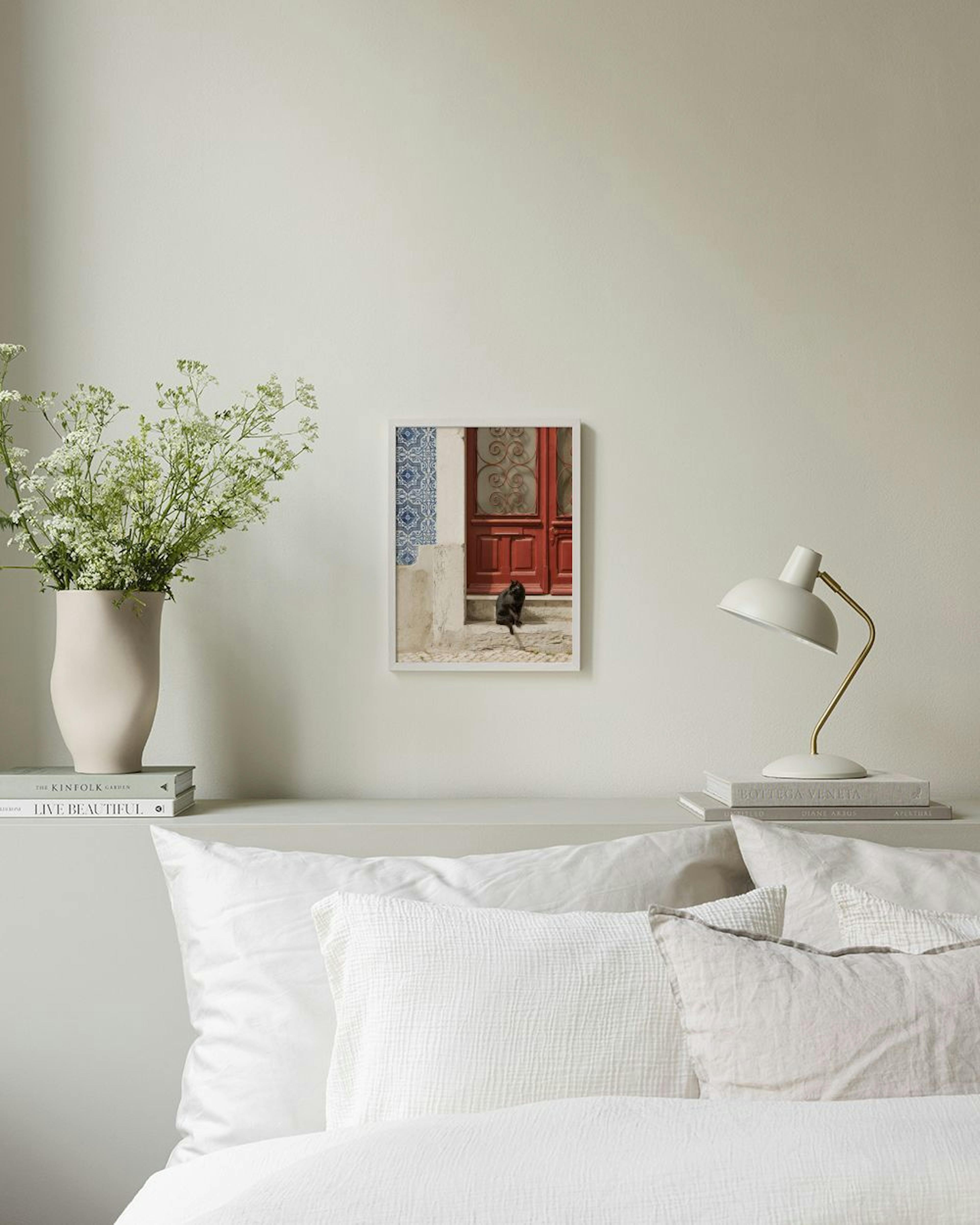 Plagát s čiernou mačkou sediacou pred červenými dverami a modrými dlaždicami, visí nad dreveným stolom s raňajkami.