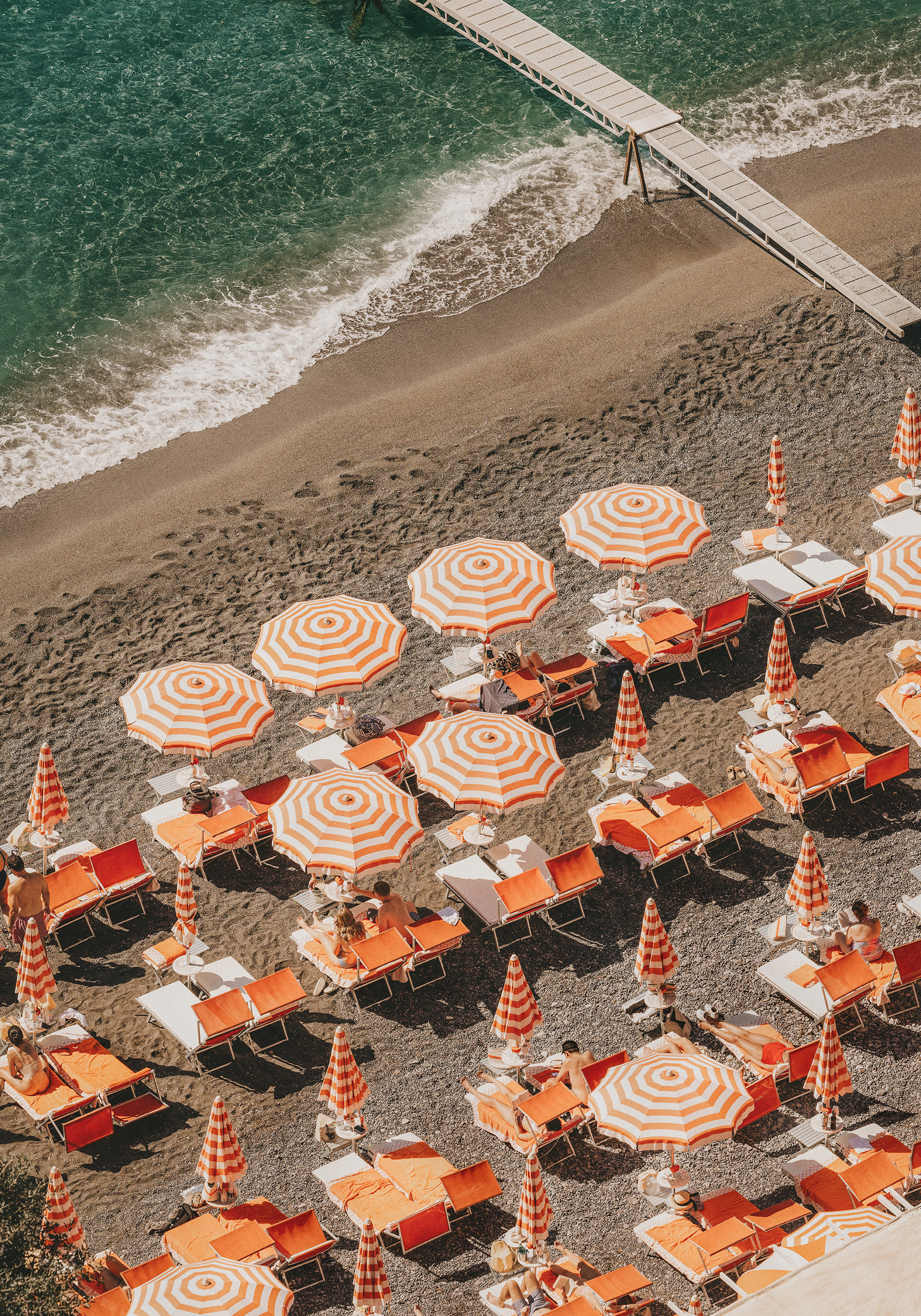 Aerial photo of Positano beach with orange and white striped umbrellas and orange lounge chairs on dark sand.
