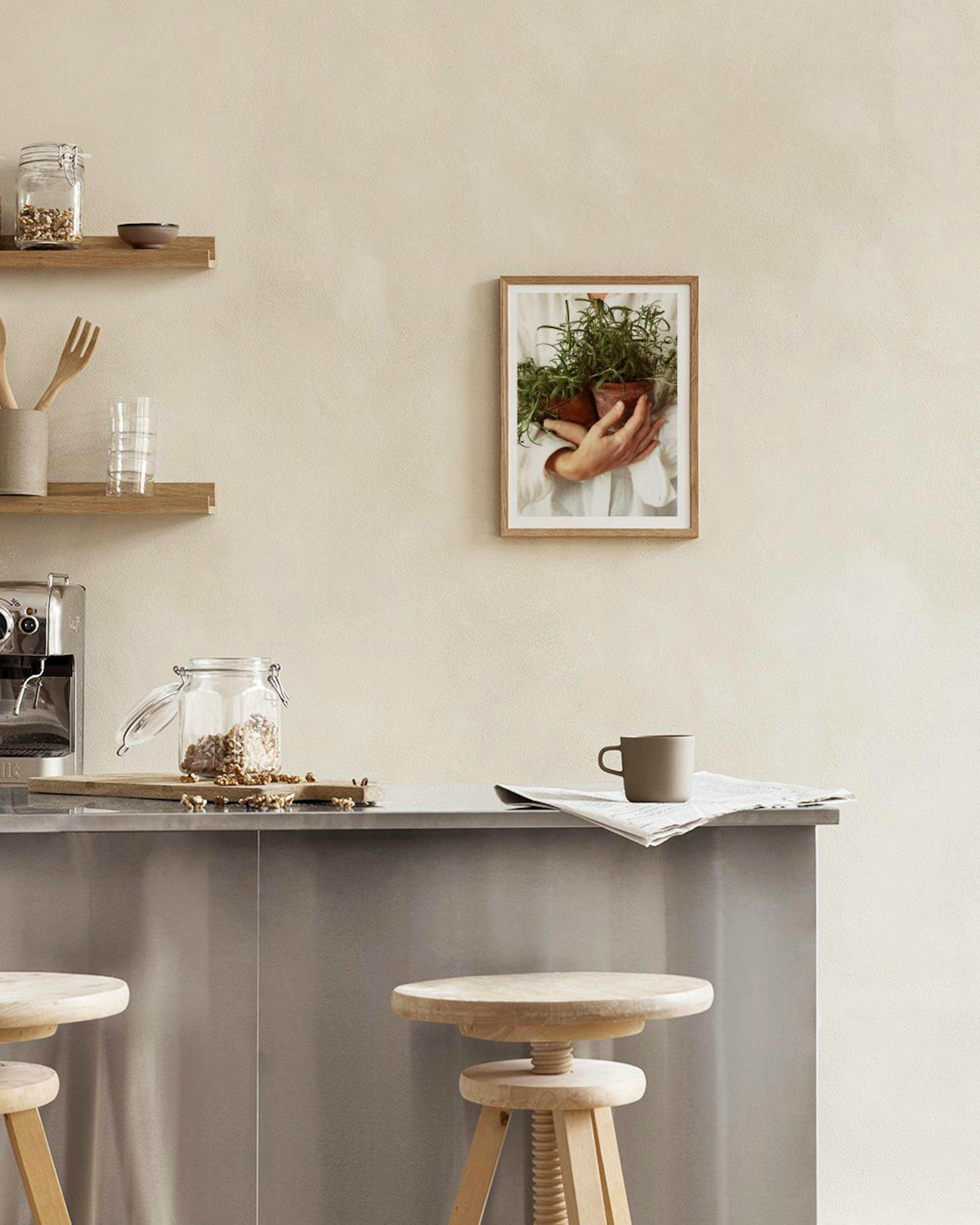 Plakat med en person, der holder to urtepotter med grønne planter, hængende i et lyst køkken over en køkkenø.