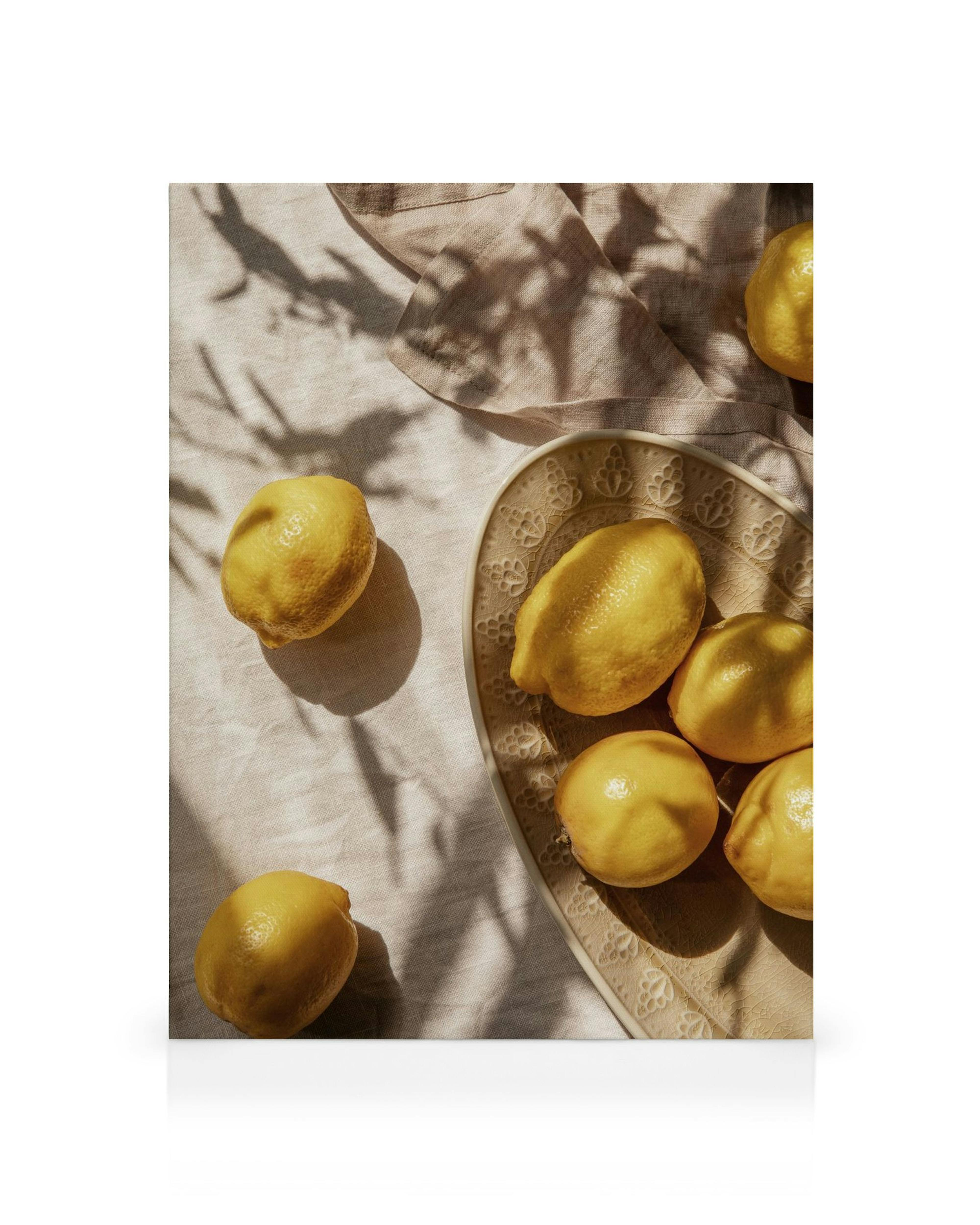 Leinwandbild mit gelben Zitronen auf einem beige gemusterten Teller, dazu Schatten von Blättern auf hellem Stoff.