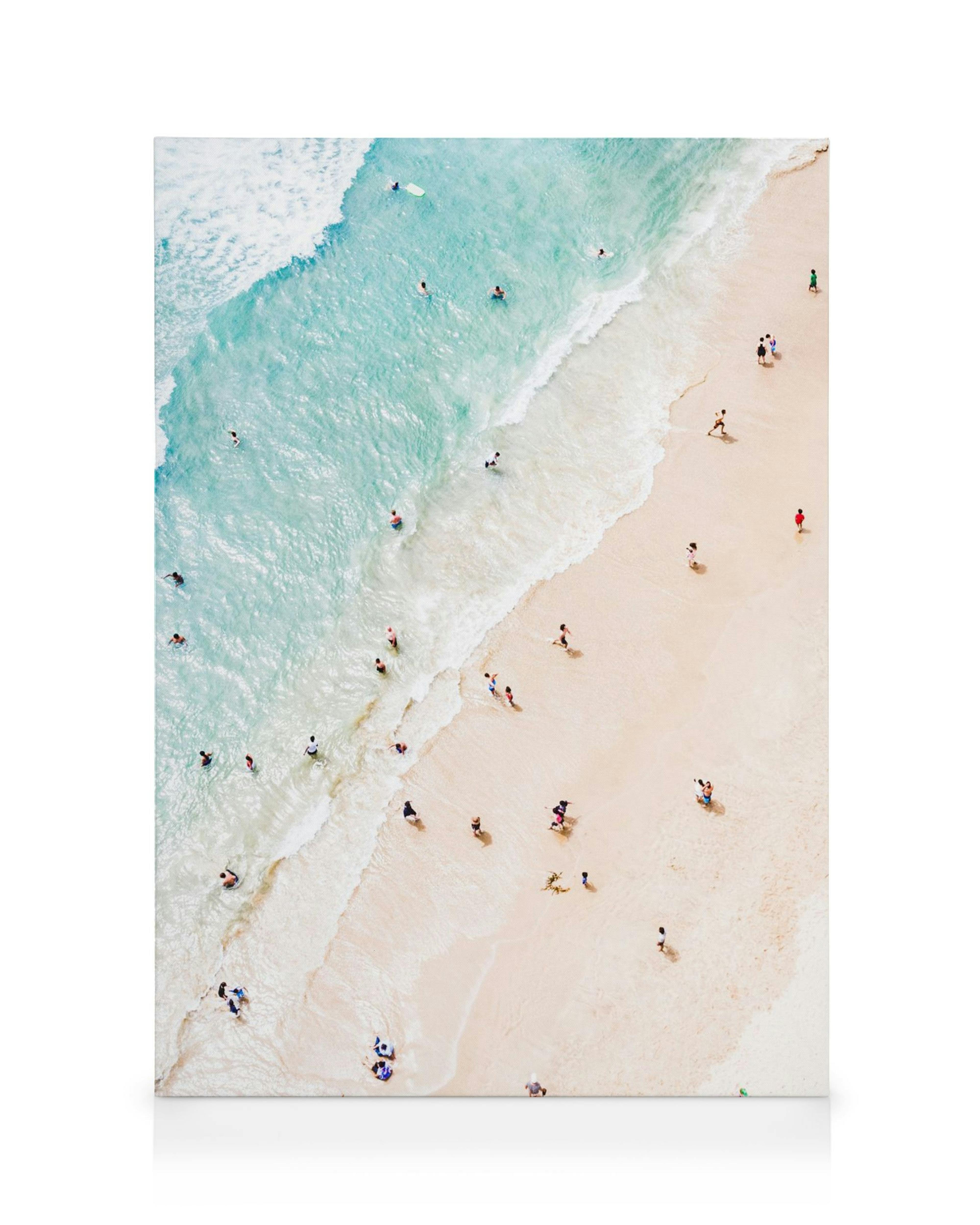 A canvas print of an aerial view of a beach with turquoise water, white waves, and people swimming and walking on the sand.