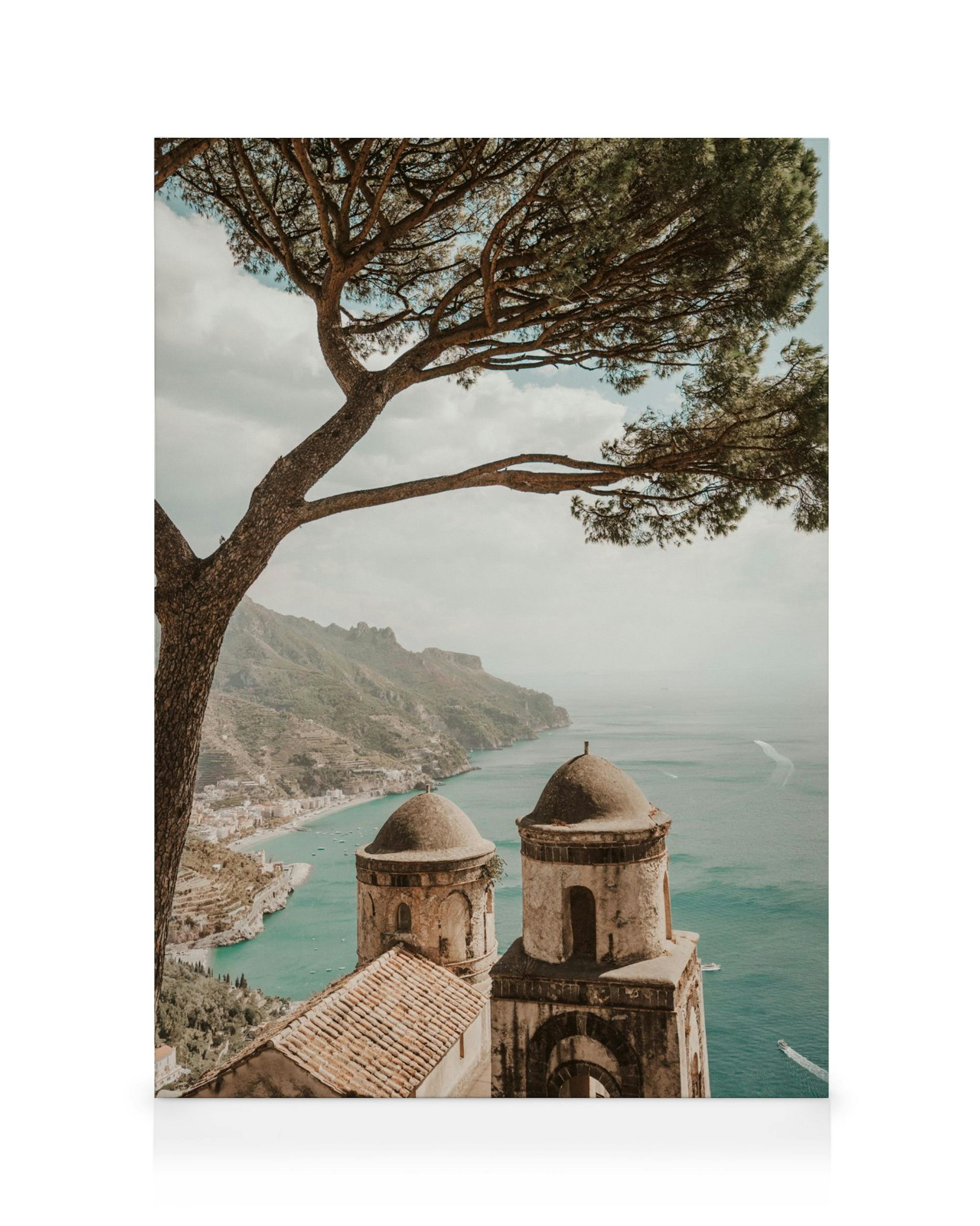 A canvas print featuring two ancient stone towers overlooking a turquoise sea and coastal town, framed by a pine tree.
