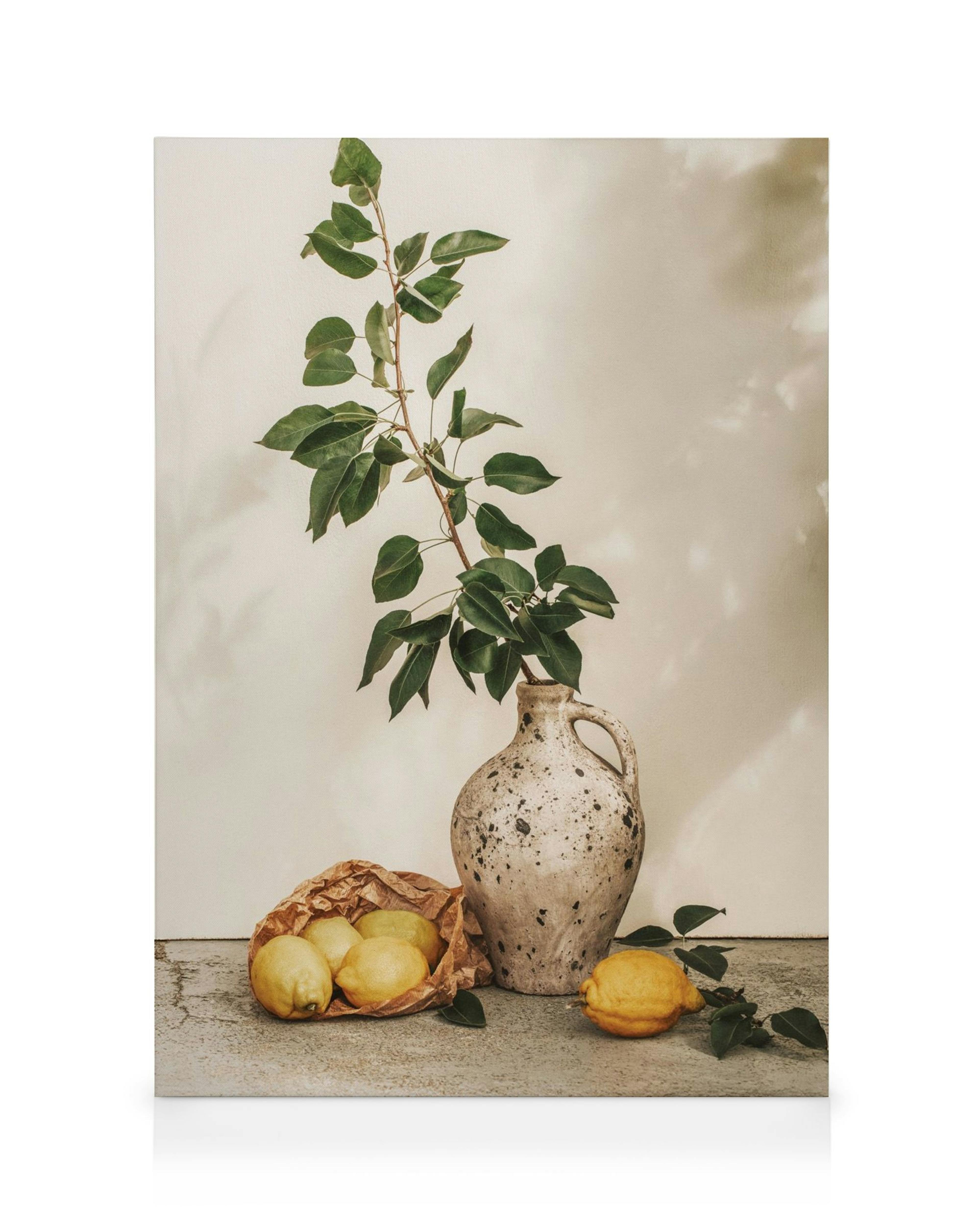A canvas print of a green branch in a rustic vase, with lemons in a paper bag and loose lemons on a concrete surface.