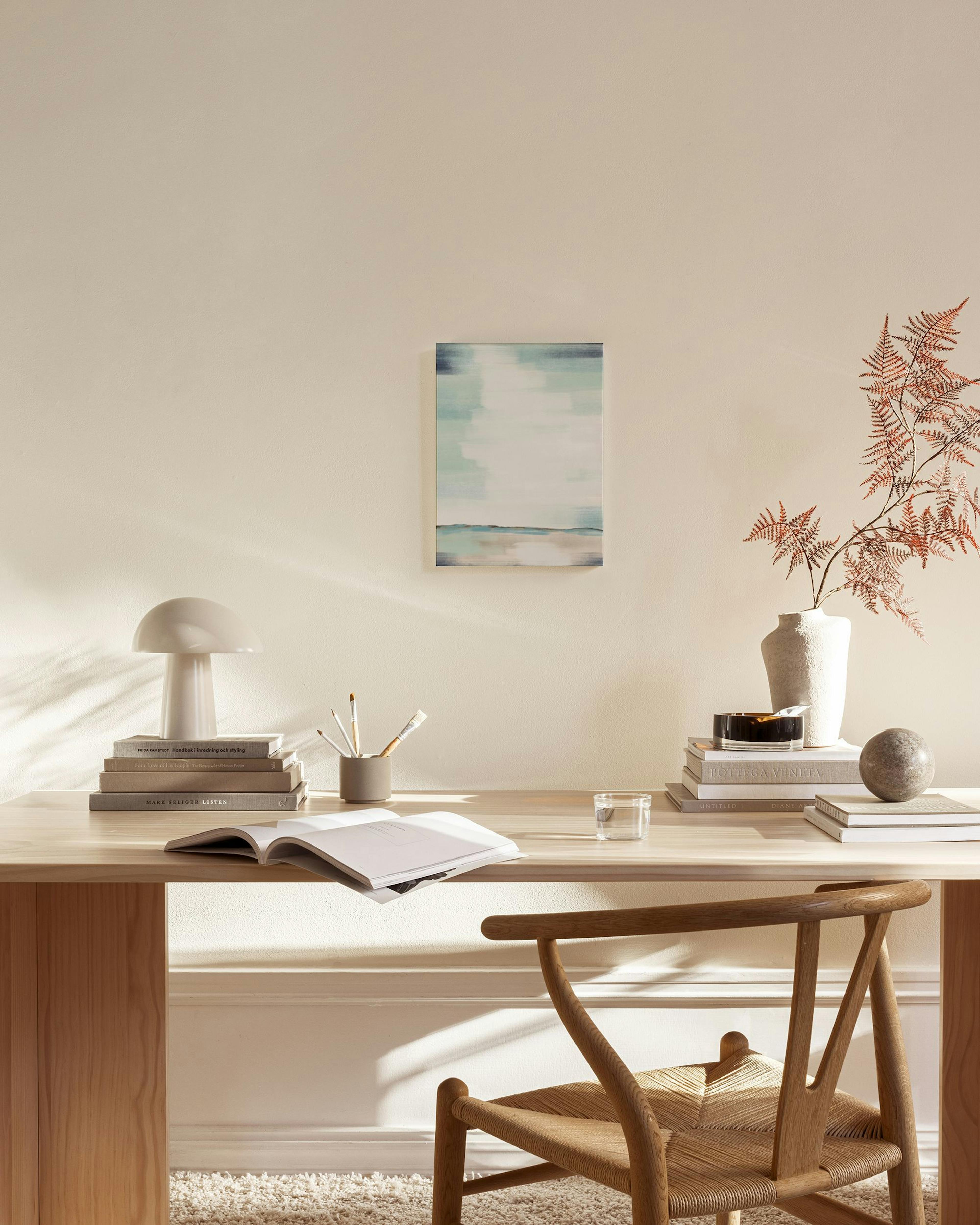 Abstraktes Leinwandbild mit horizontalen Pinselstrichen in Blau, Türkis und Beige, an heller Wand über einem Holztisch.