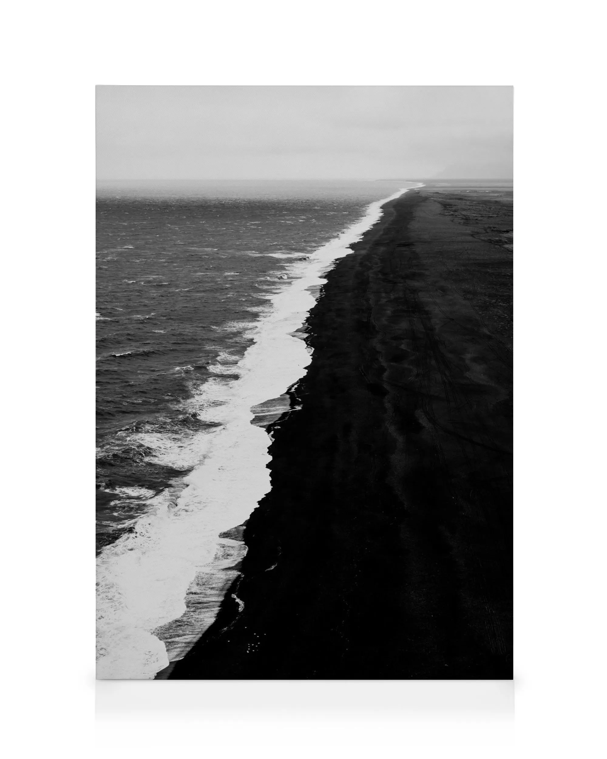 Aerial black and white canvas of a black sand beach with white ocean waves crashing along the shore under a pale sky.