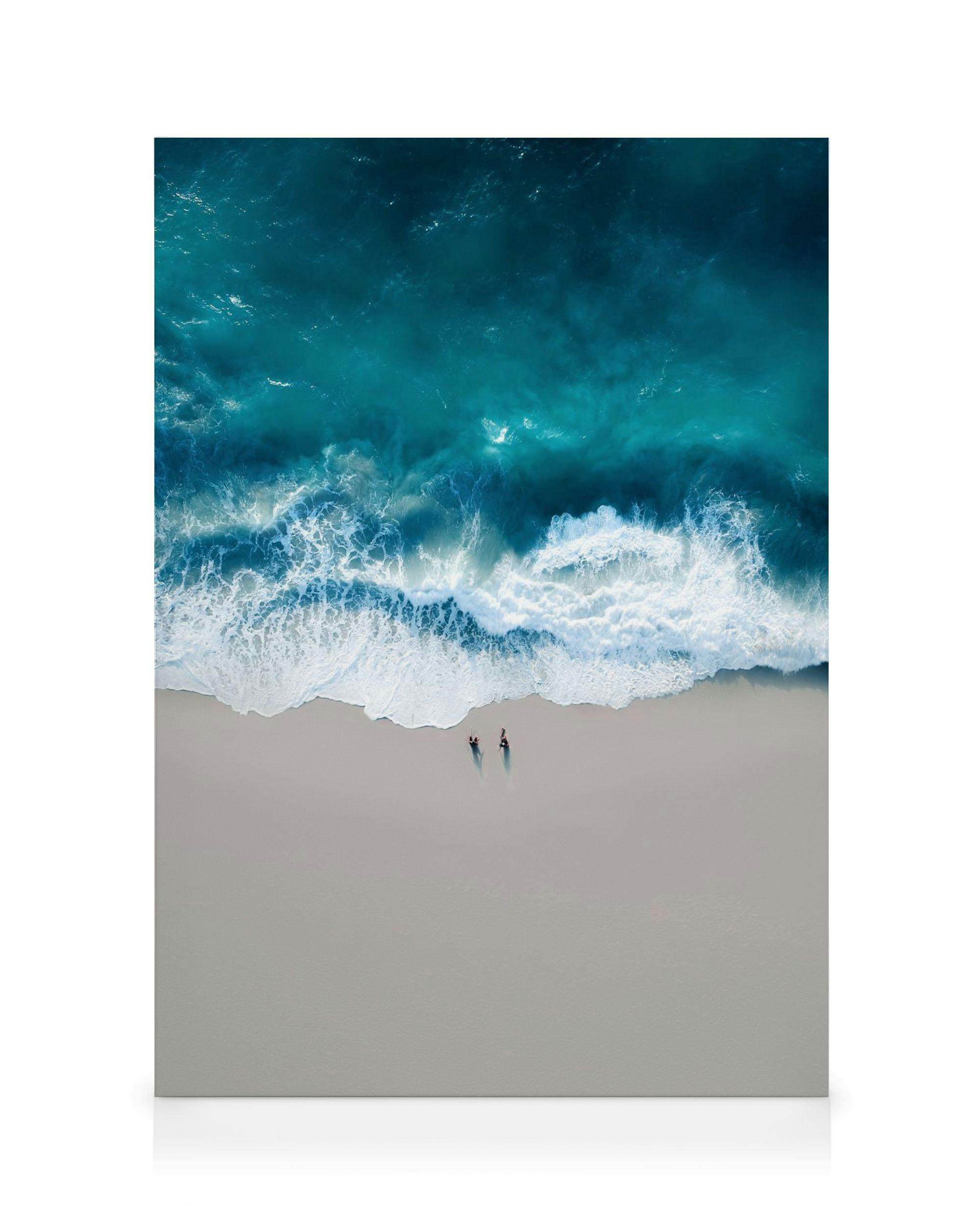 Aerial view of turquoise ocean waves crashing onto a sandy beach with two tiny figures walking along the shore