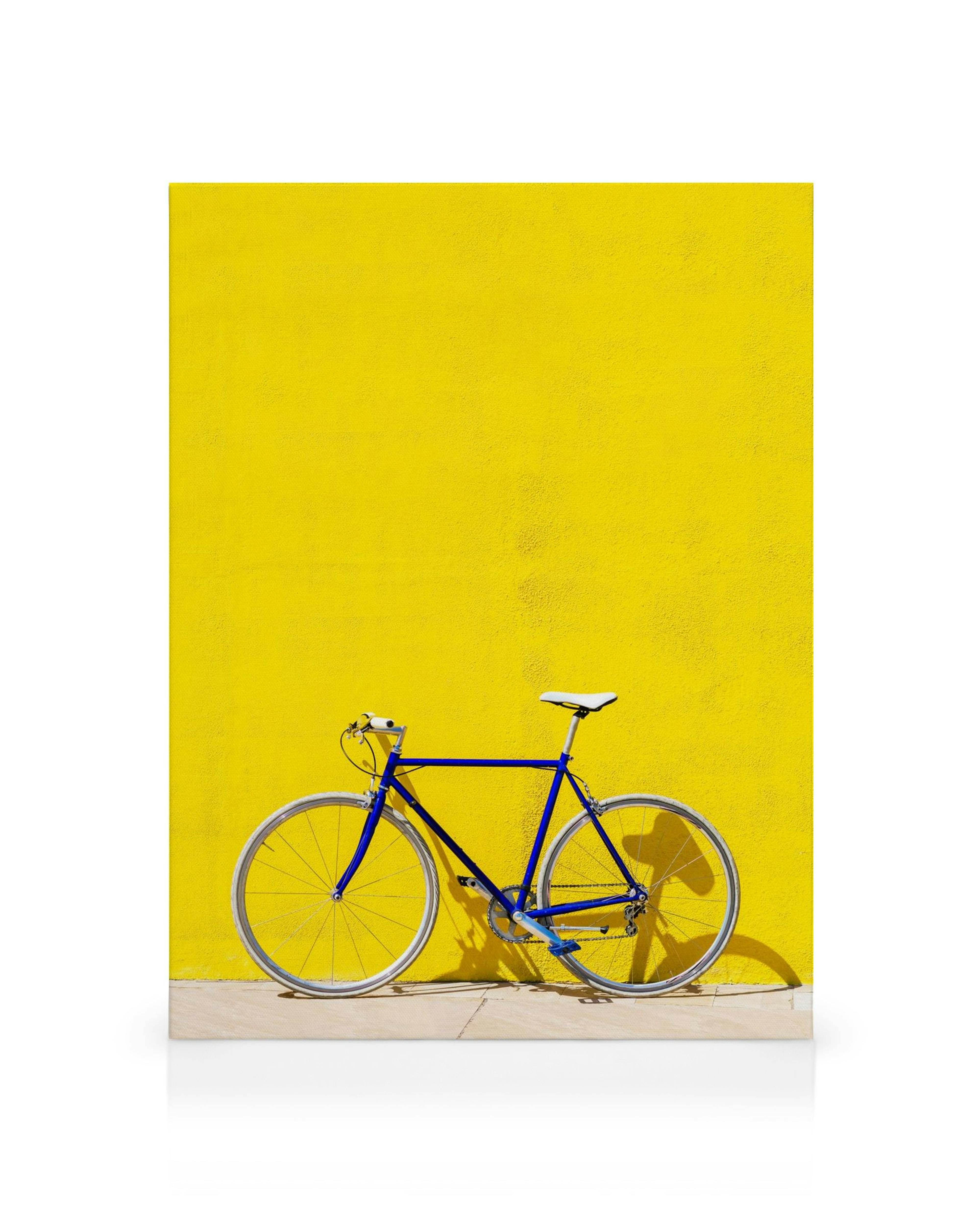 Lienzo de bicicleta azul con neumáticos blancos contra una pared amarilla brillante y texturizada, con sombra en el suelo gris.