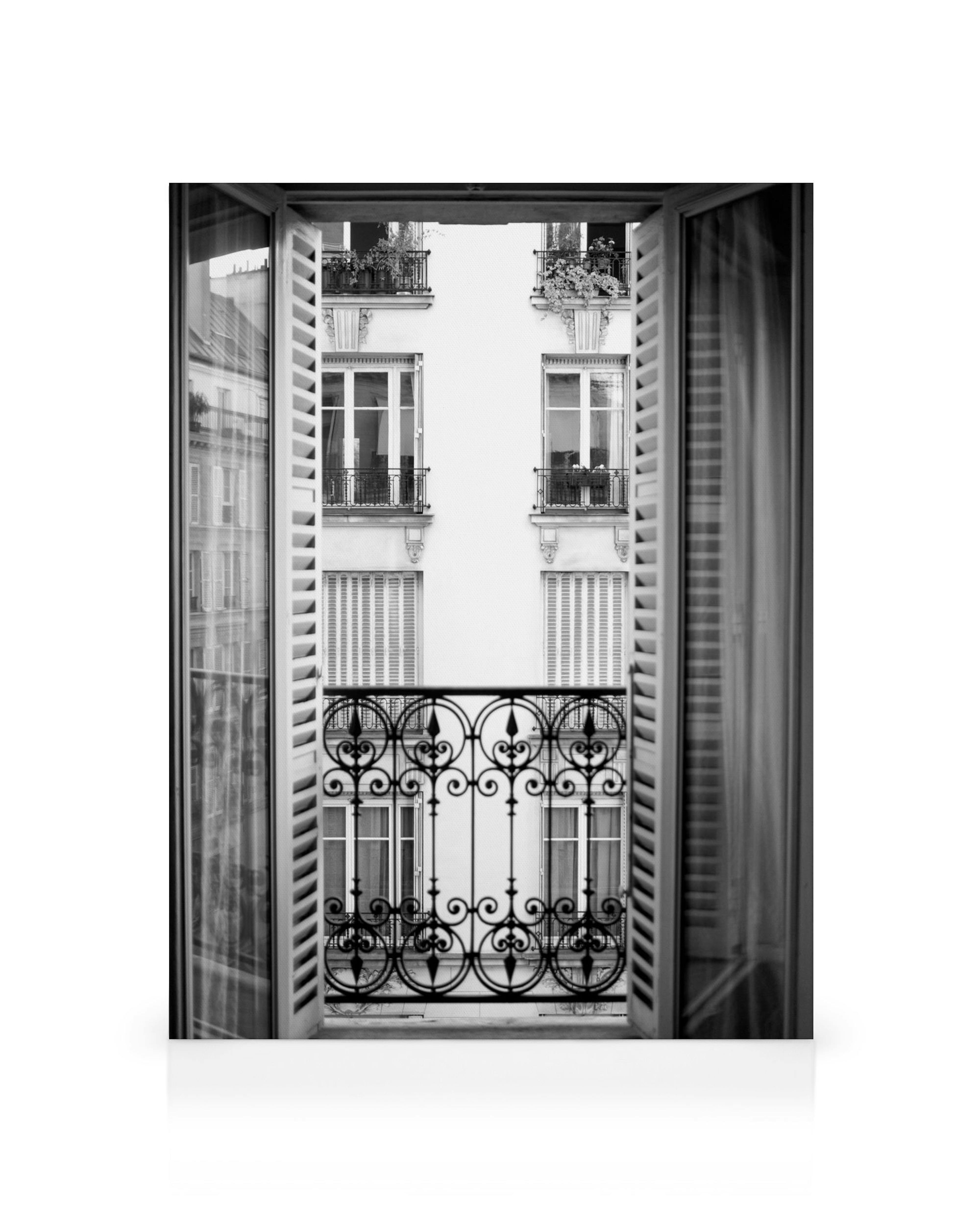 Fönsterutsikt i svartvitt över parisisk byggnad med balkongräcke i smidesjärn och öppna fönsterluckor.