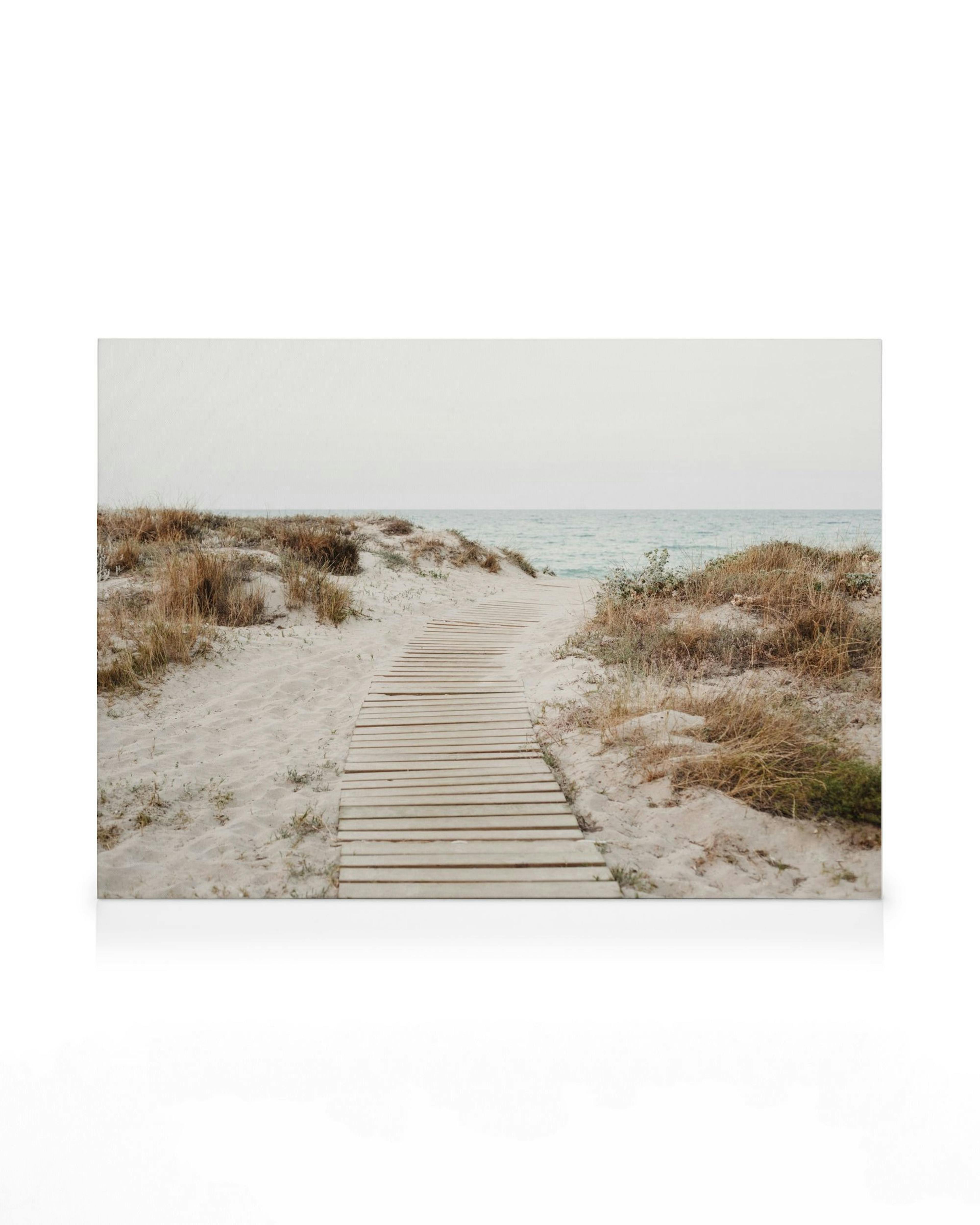 Toile de plage, chemin en bois dans le sable vers la mer, herbe, tons blancs et bleus, ciel couvert. Typographie