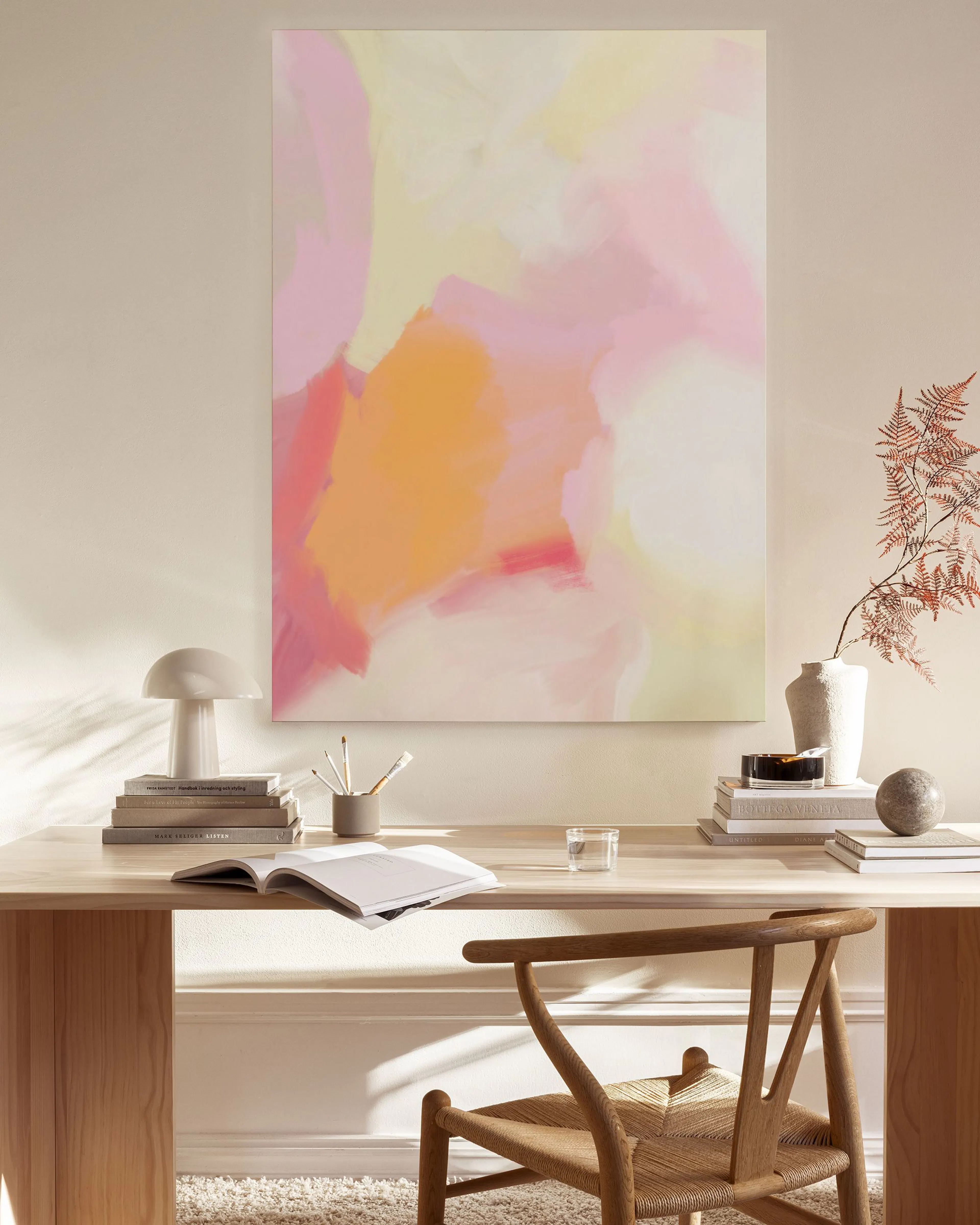 Toile abstraite aux tons rose, orange et jaune, accrochée dans un salon lumineux avec fauteuil en bois et table basse.