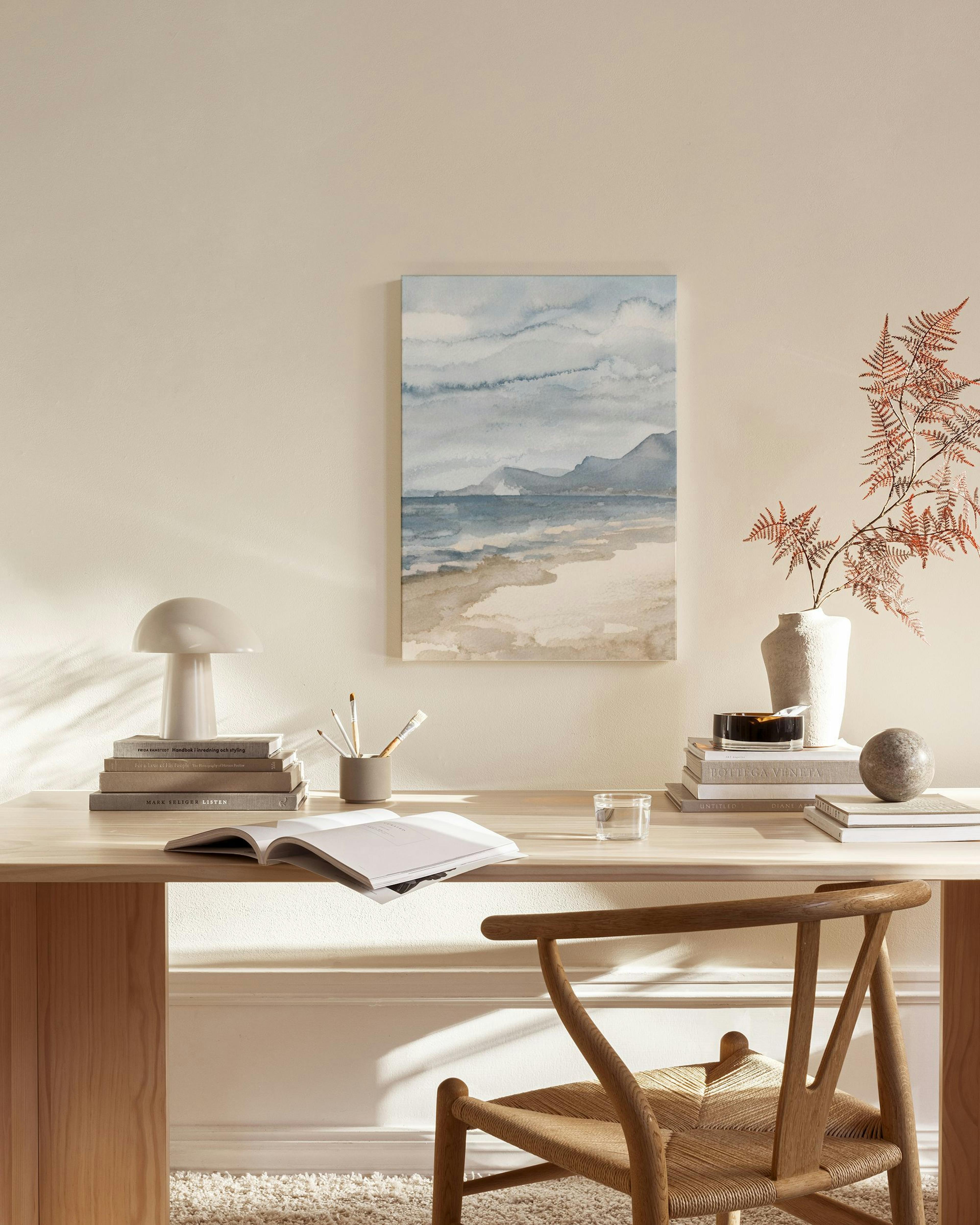 Canvastavla med akvarellmålning av ett kustlandskap med hav, strand och berg i blå och bruna toner, hängandes i ett ljust vardag