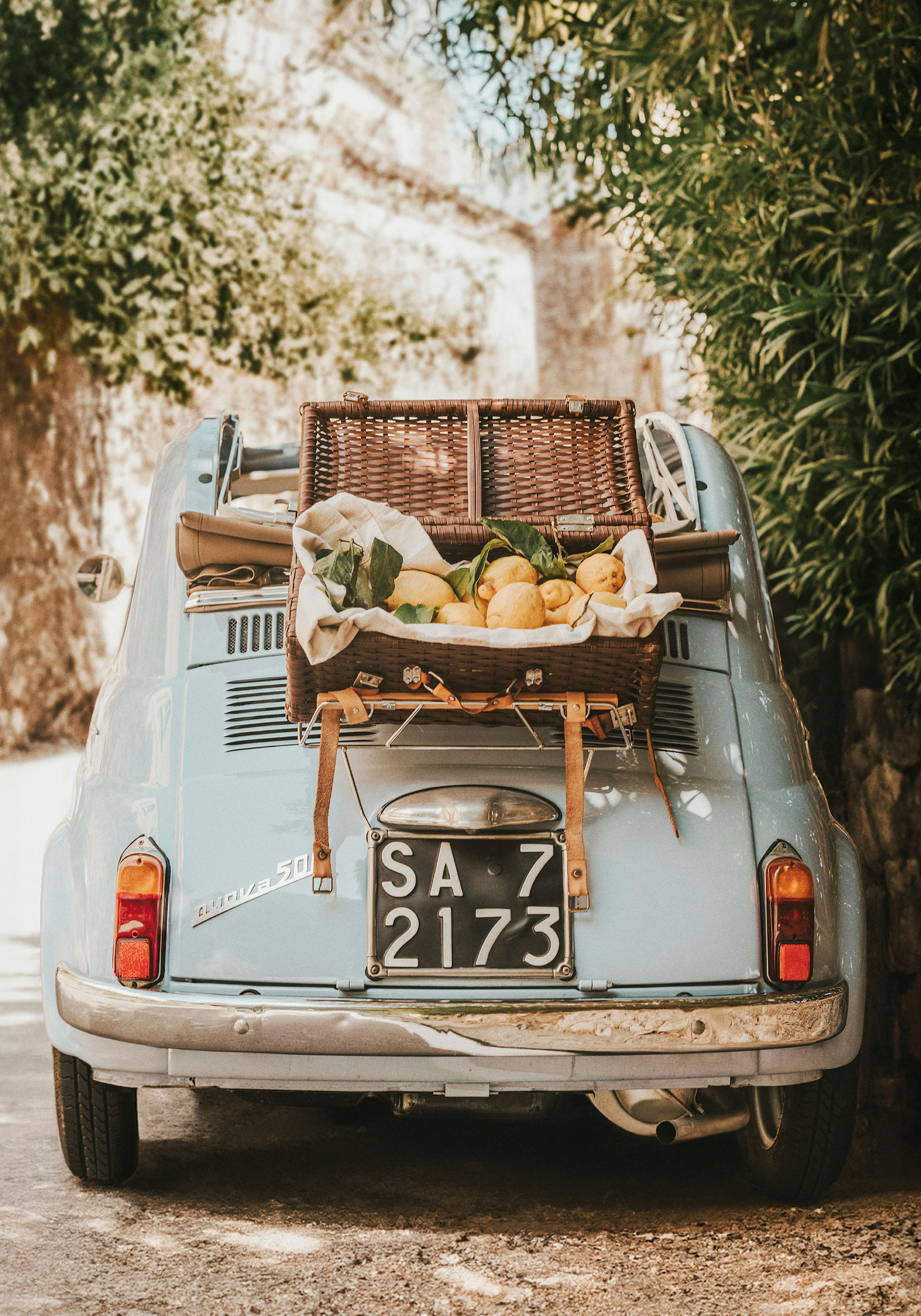 Ρετρό μπλε αυτοκίνητο με καλάθι πικνίκ, γεμάτο λεμόνια, σε ιταλικό τοπίο.