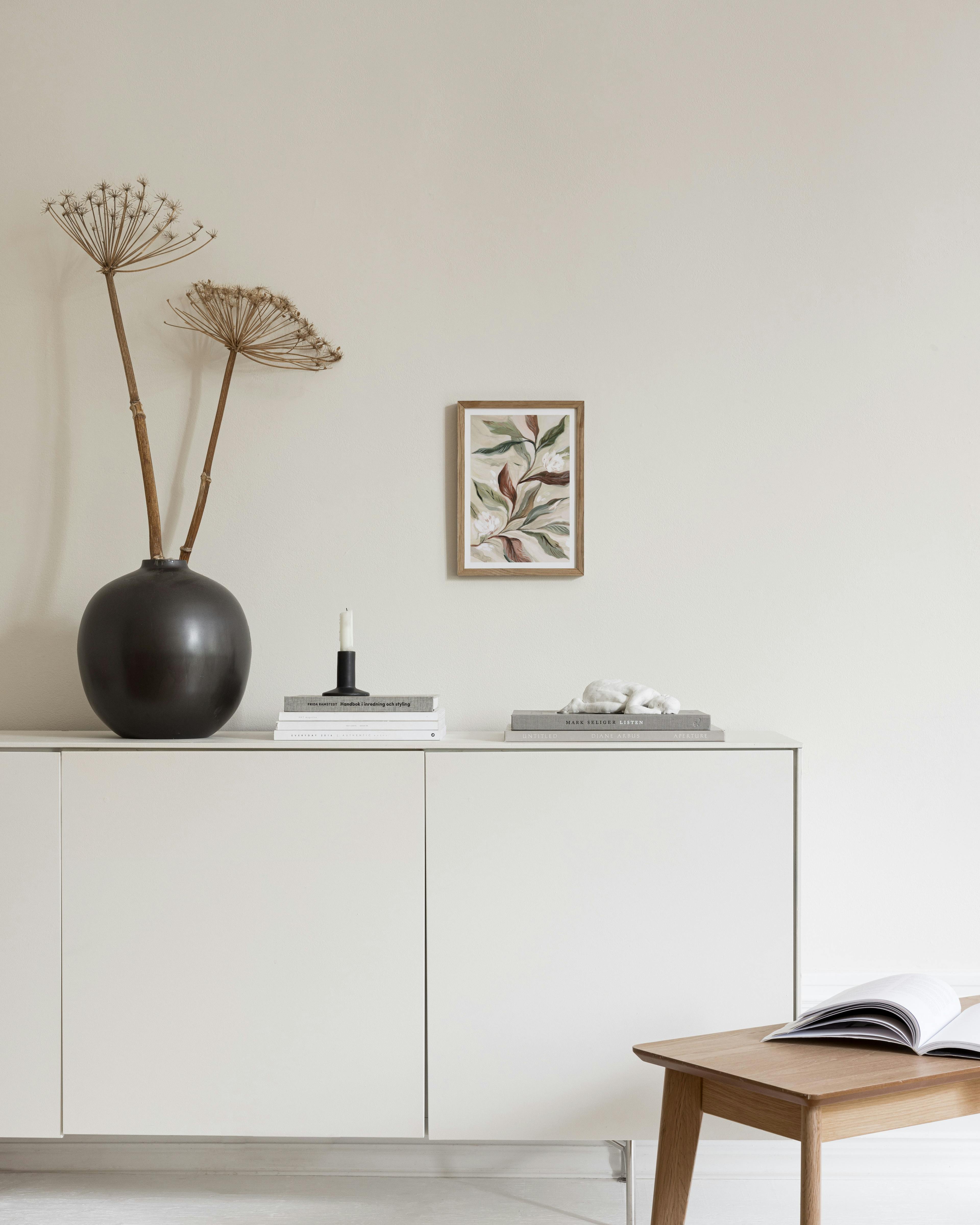 A botanical print with white flowers and green and brown leaves, hanging above a white console table with a black vase and books