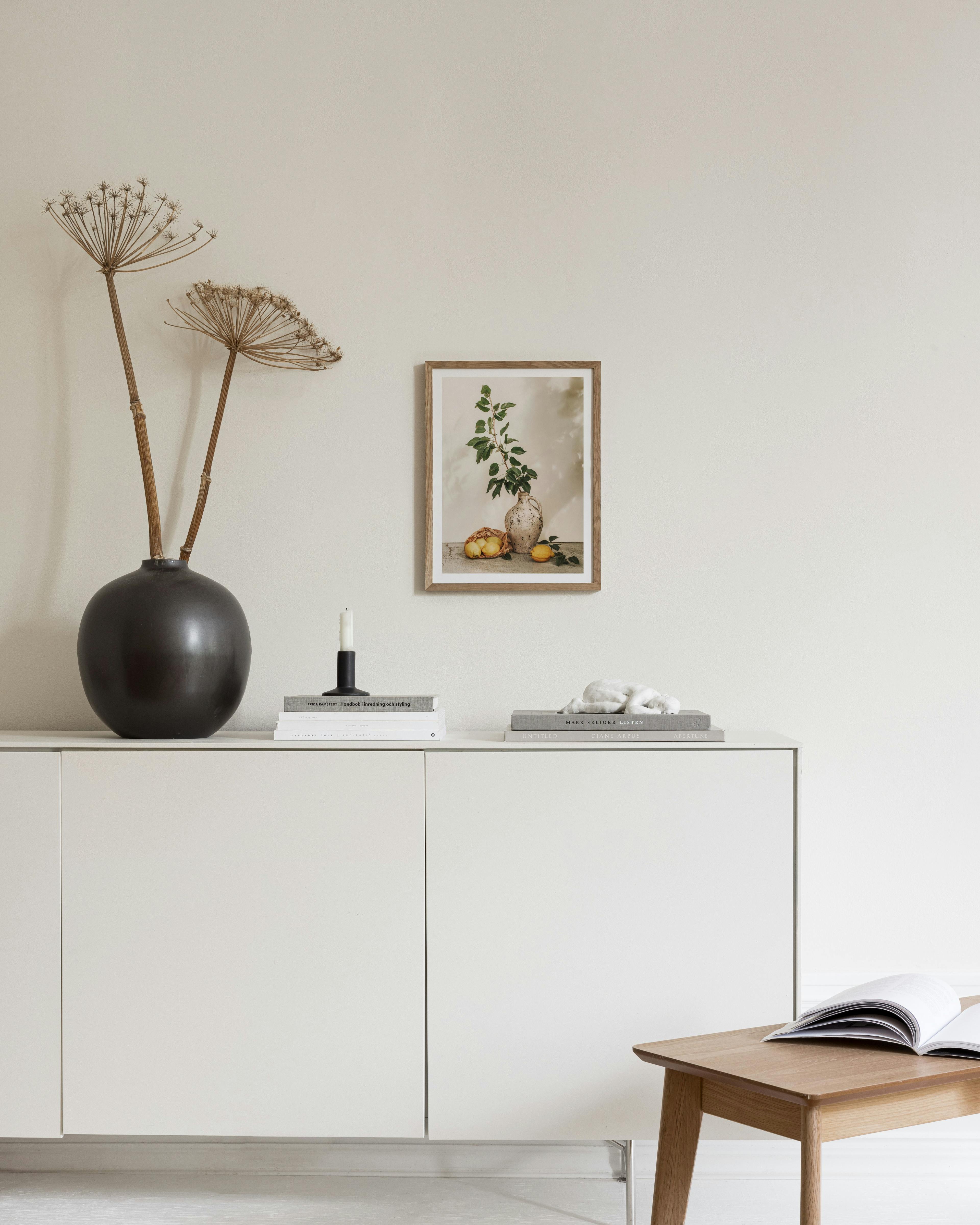 Plakat med citroner og en gren i vase, hængende over et hvidt skab i et lyst rum.