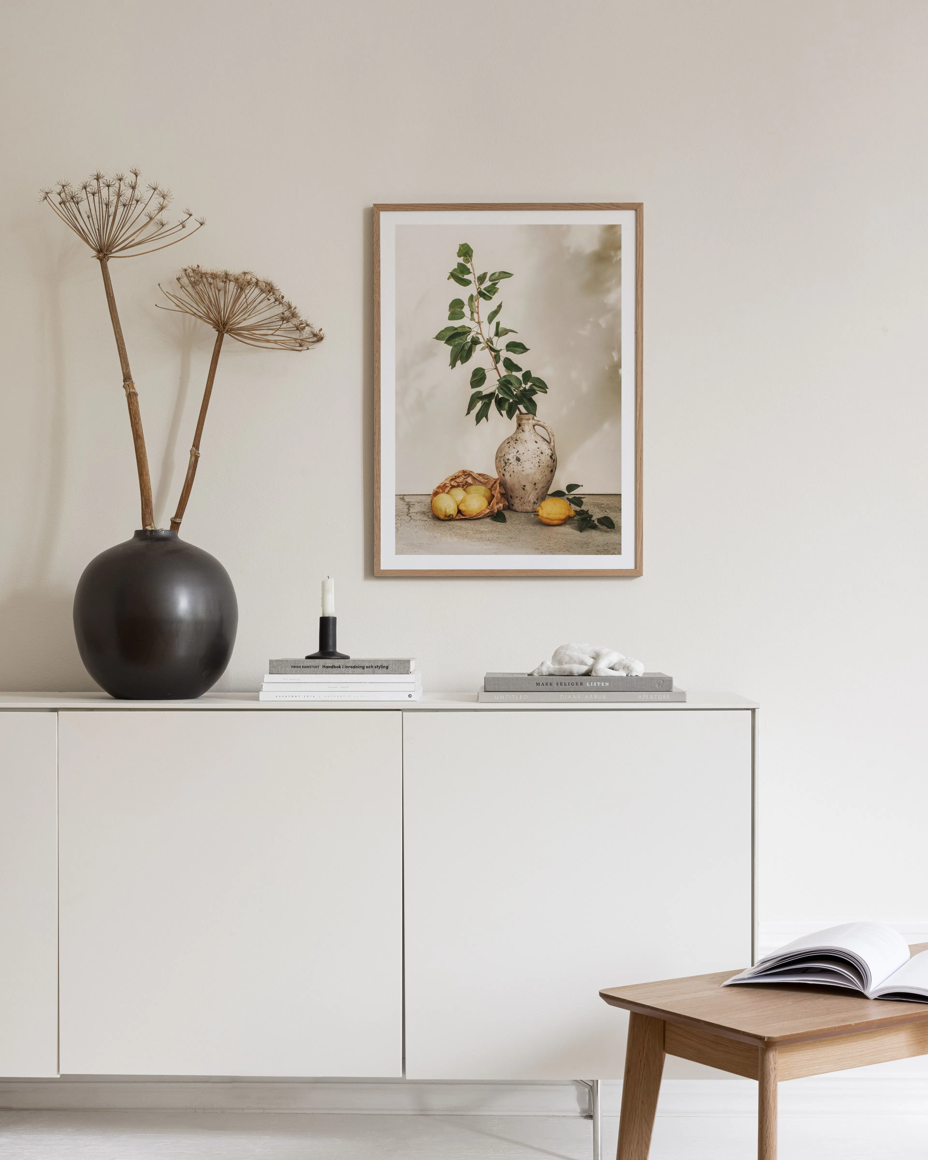 En poster med en vas med gröna blad och citroner, hängande ovanför ett vitt skåp med inredningsdetaljer.