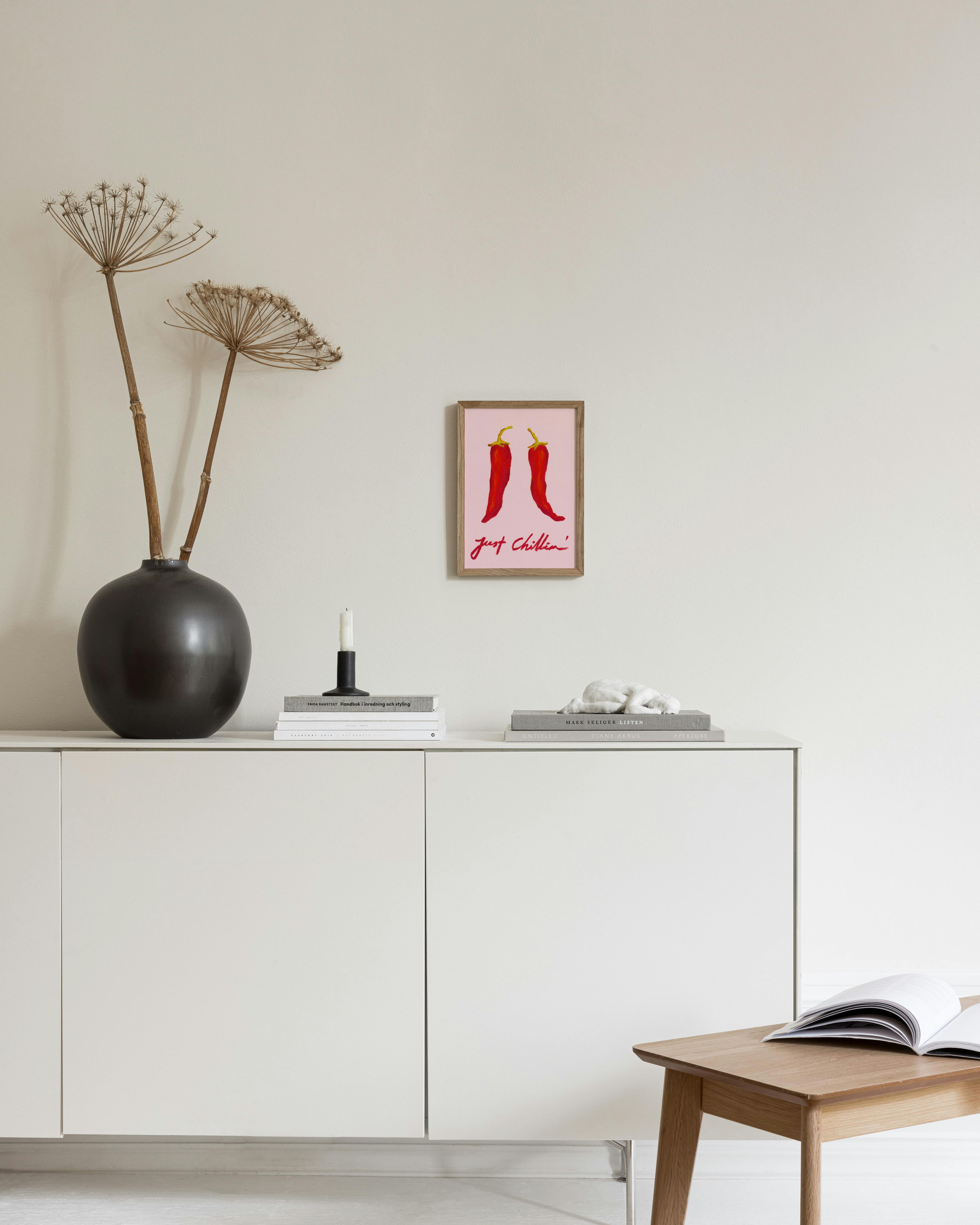 Juliste kahdesta punaisesta chilipaprikasta vaaleanpunaisella pohjalla ja tekstillä just Chillin, valkoisen kaapin yläpuolella.