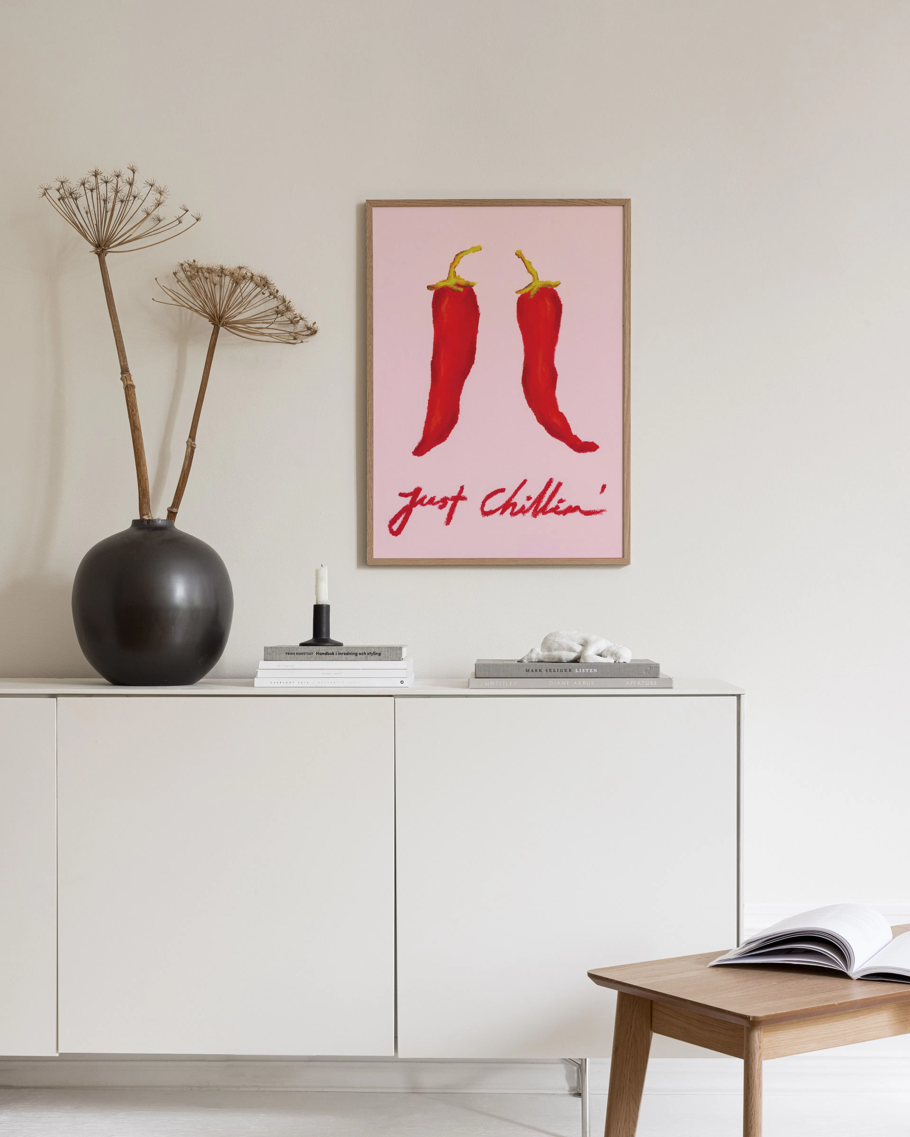 Póster de dos chiles rojos sobre fondo rosa con la frase just chillin, colgado sobre un mueble blanco en una sala moderna.