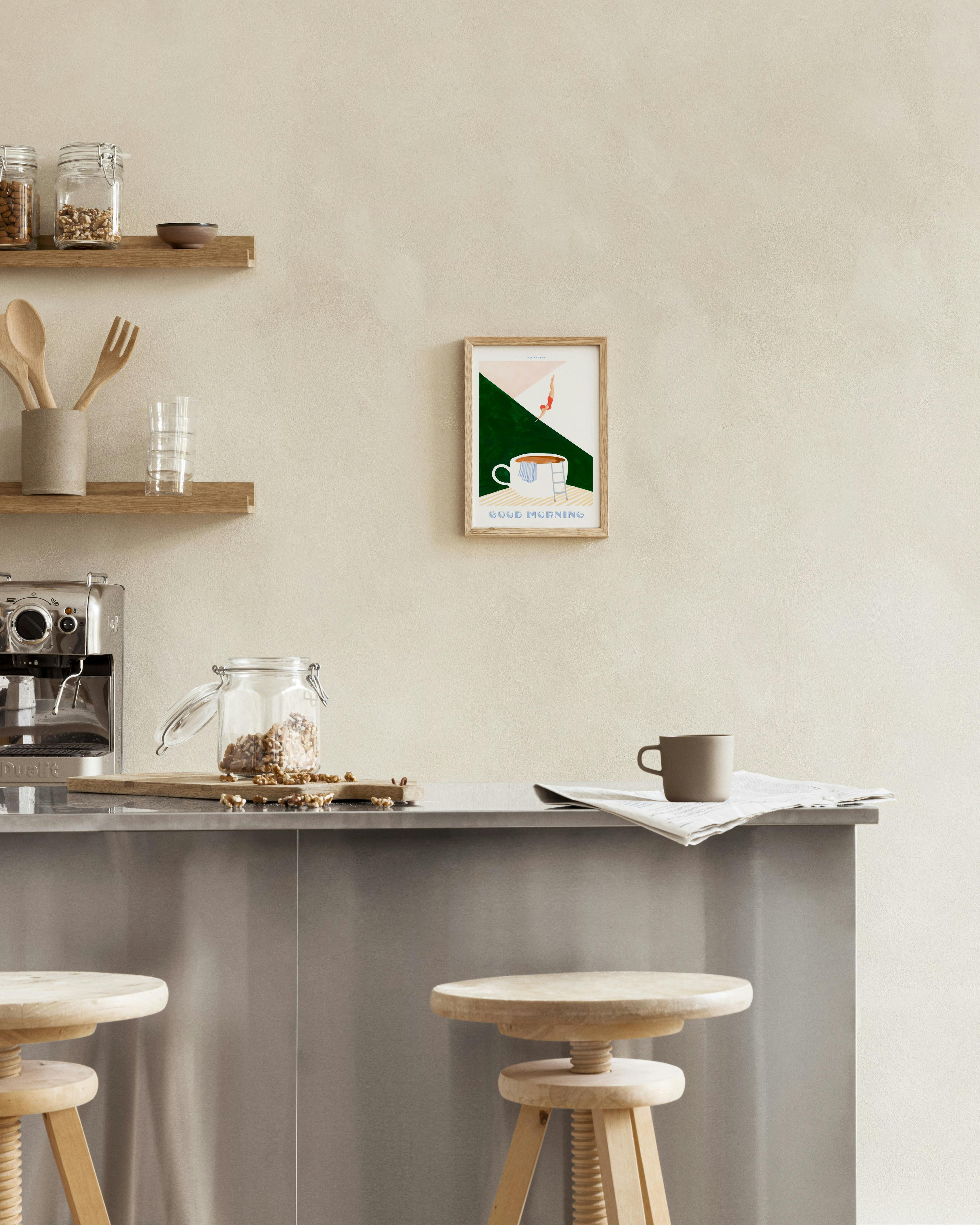 Poster Good Morning“ mit einer Frau beim Sprung in eine Kaffeetasse, an einer Wand über einem Holztisch.