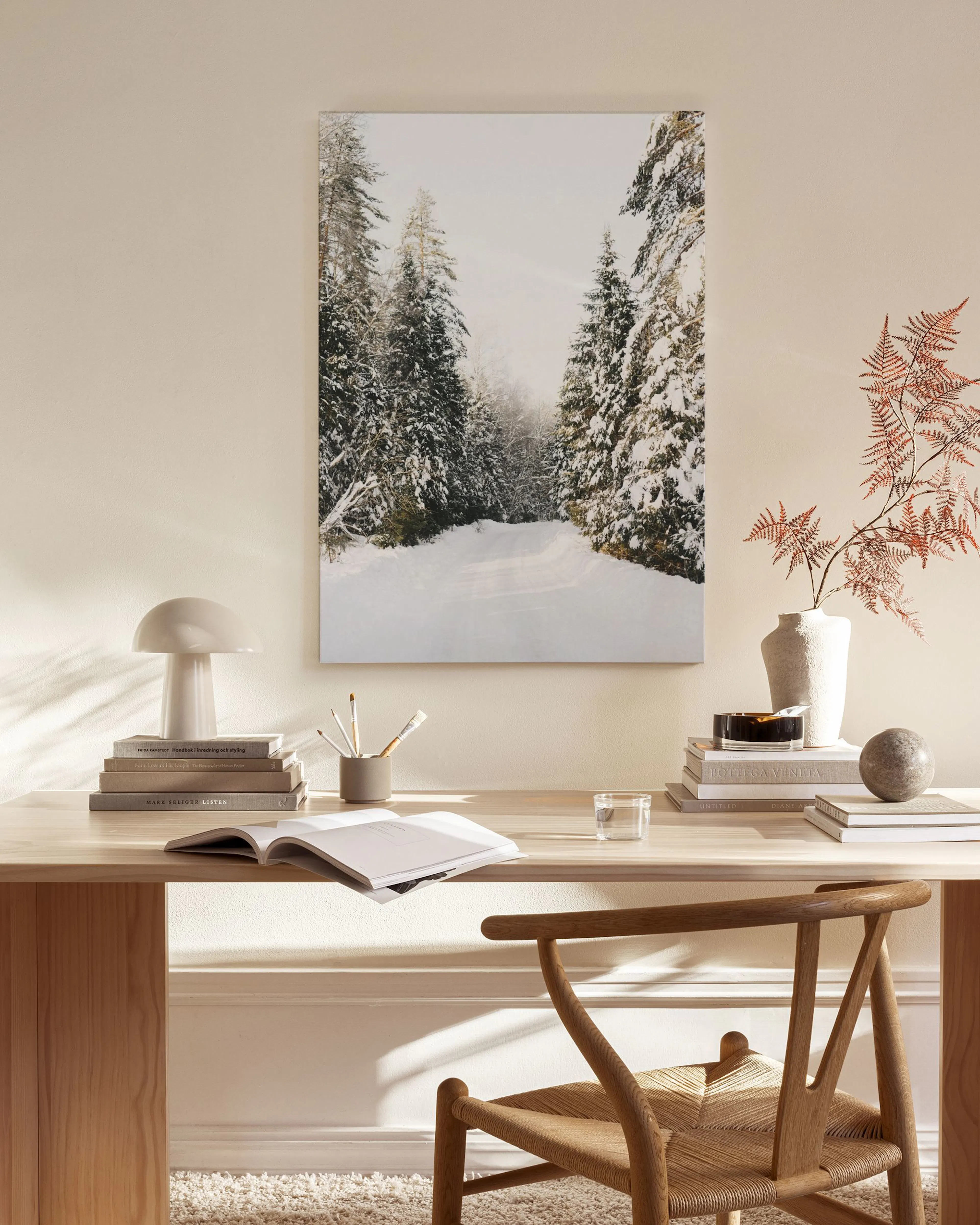 A canvas print of a snowy forest road with tall evergreen trees, displayed in a bright living room above a wooden console table.