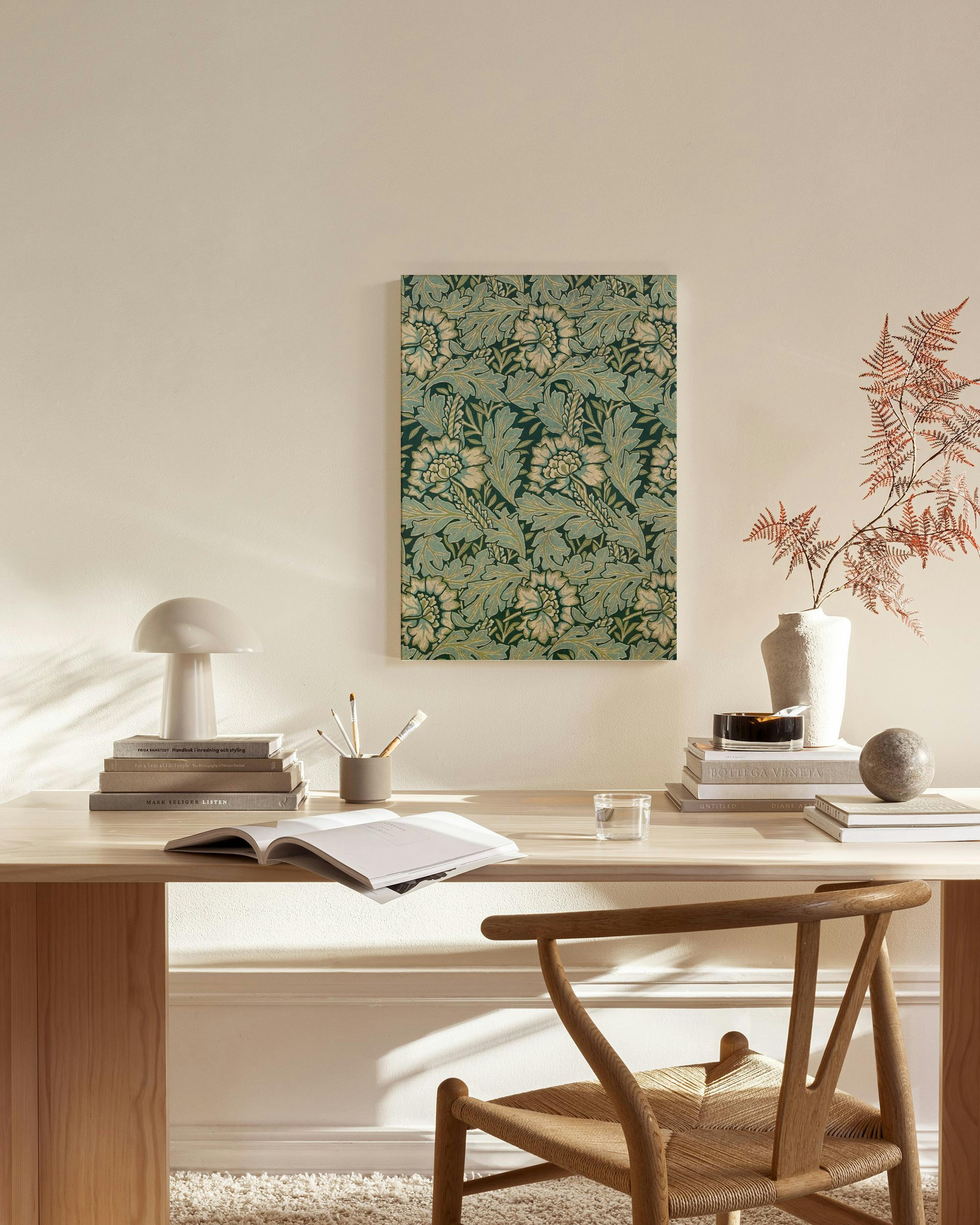 Καμβάς με επαναλαμβανόμενο μοτίβο από μπεζ και πράσινα φύλλα και λουλούδια, κρεμασμένος σε μπεζ τοίχο πάνω από ξύλινο έπιπλο.