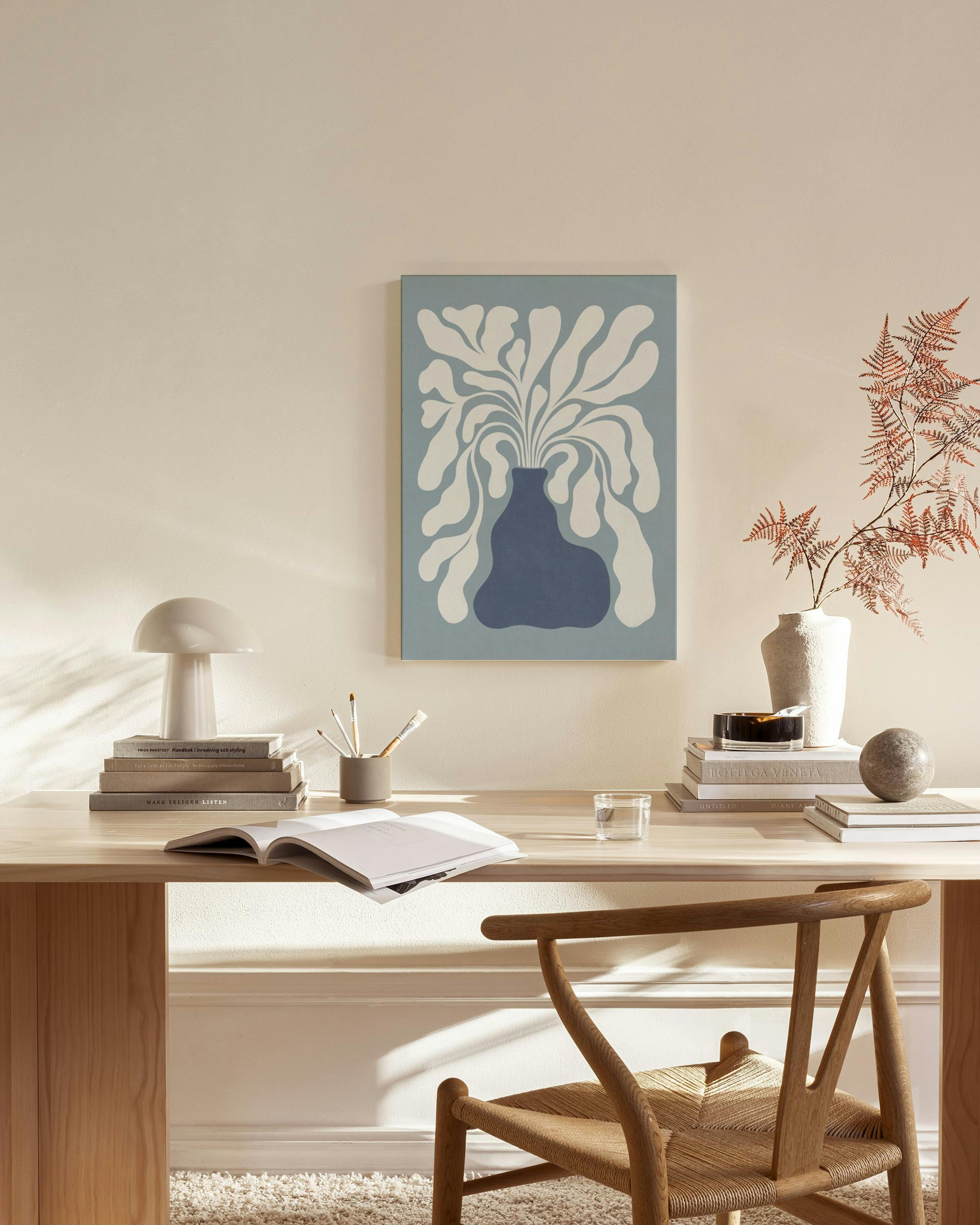 Obraz na płótnie z wazonem w odcieniach błękitu i białymi liśćmi, wiszący w salonie nad komodą obok fotela.