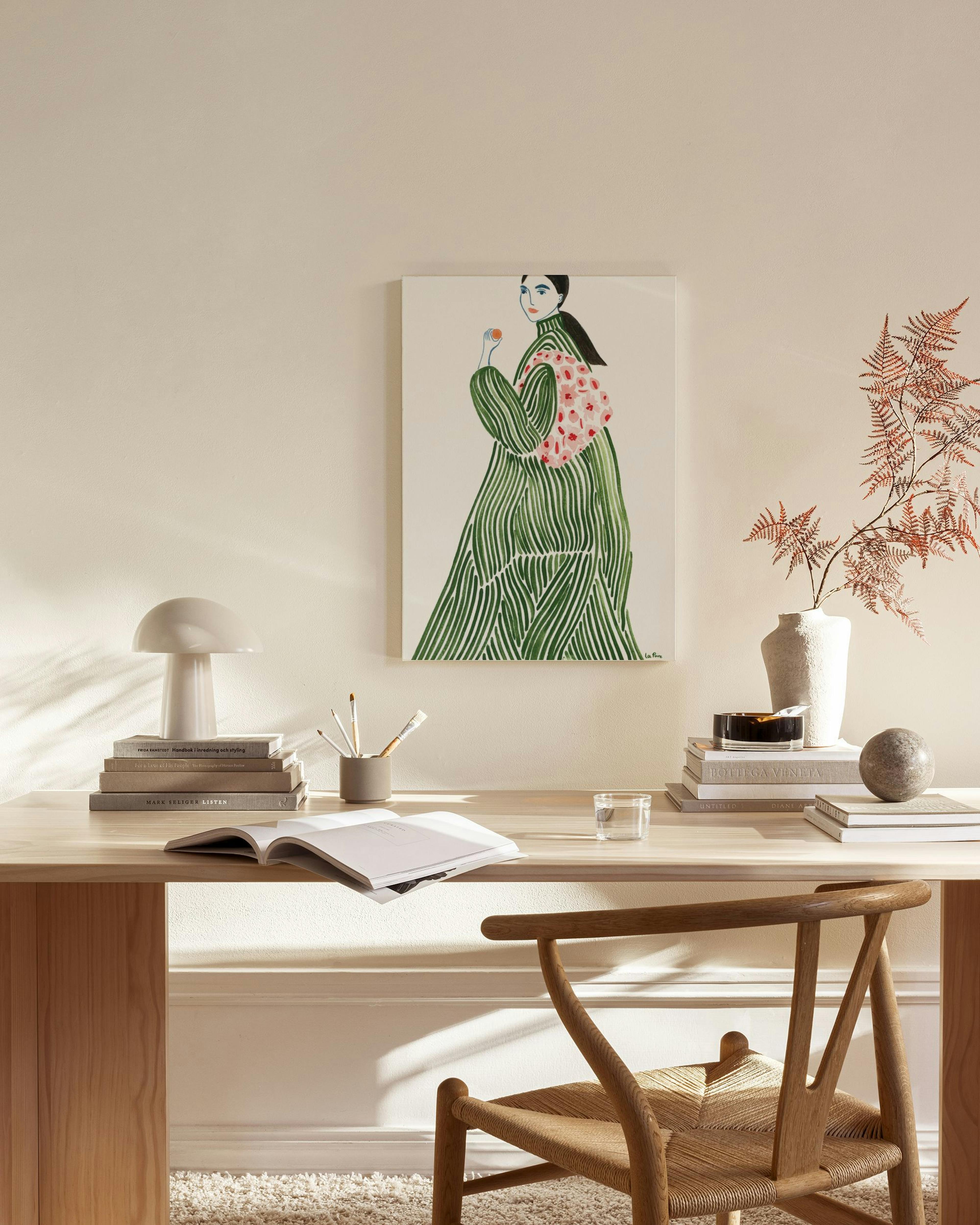 Καμβάς με εικονογράφηση γυναίκας με πράσινο ριγέ φόρεμα και ροζ φουλάρι, κρεμασμένος σε μπεζ τοίχο πάνω από ξύλινο έπιπλο.