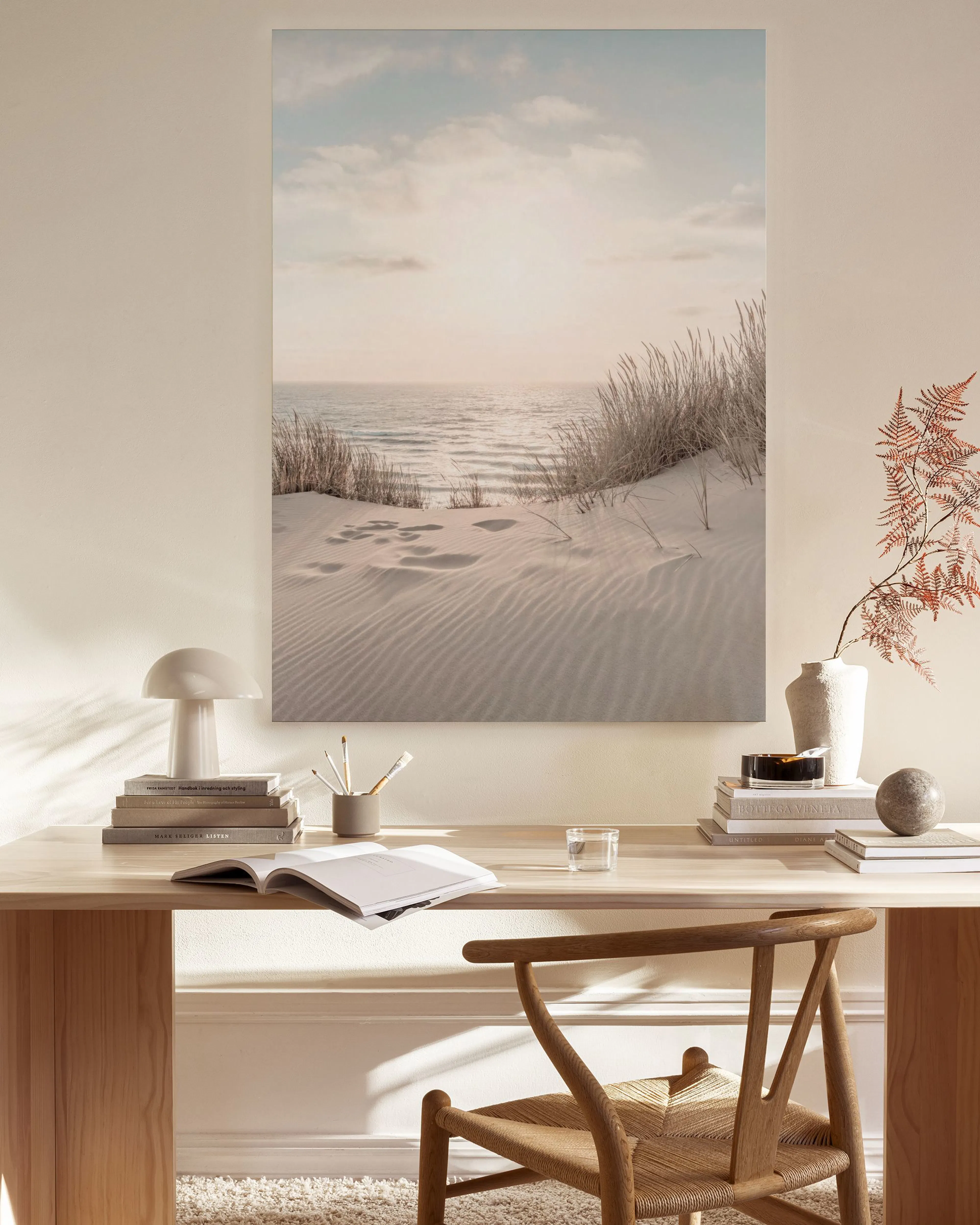 A canvas print of a sandy beach with footprints and tall grass at sunset, hanging above a wooden console table in a minimalist l