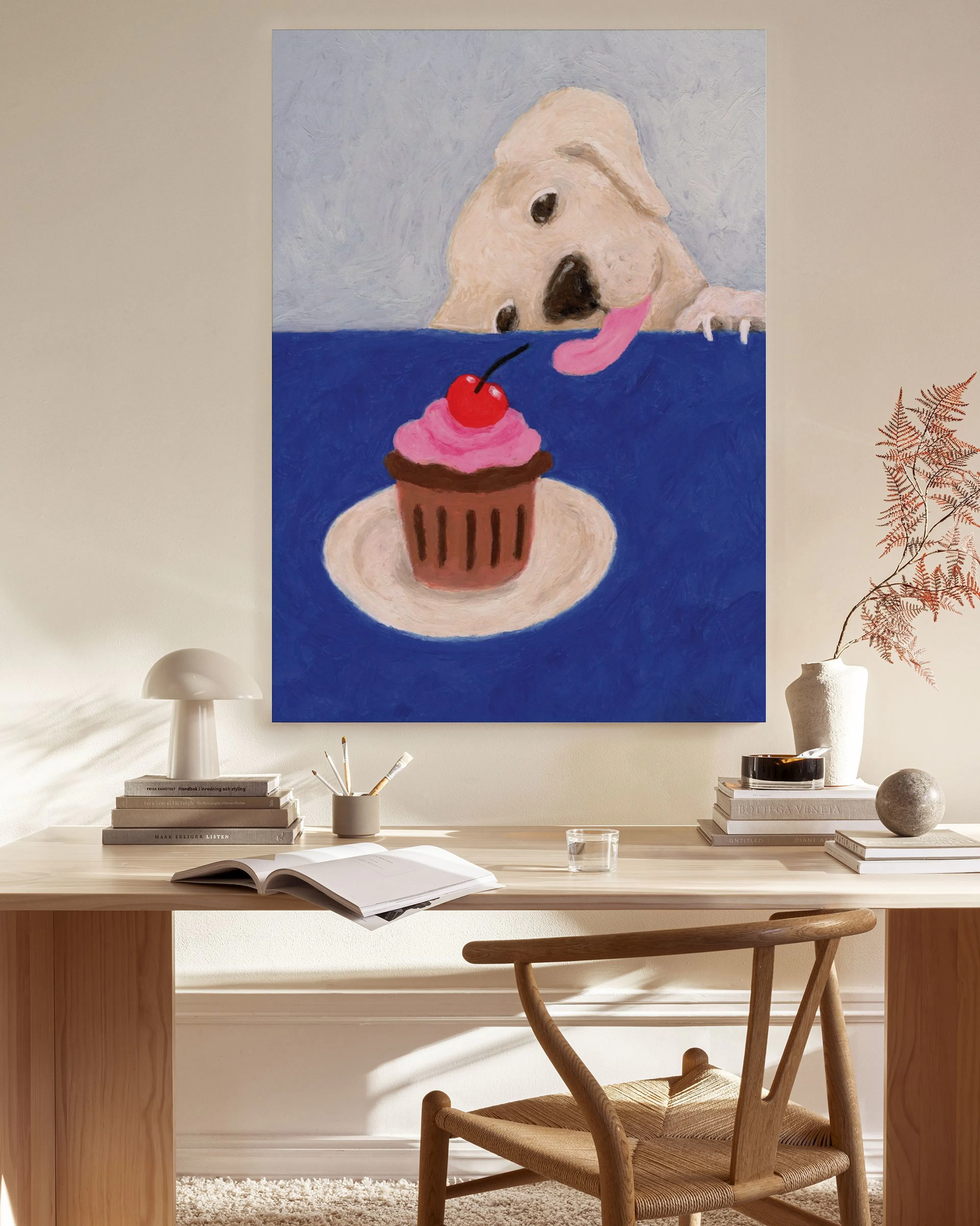Lienzo de un perro beige con la lengua afuera, mirando un cupcake con cereza, colgado en una sala moderna.