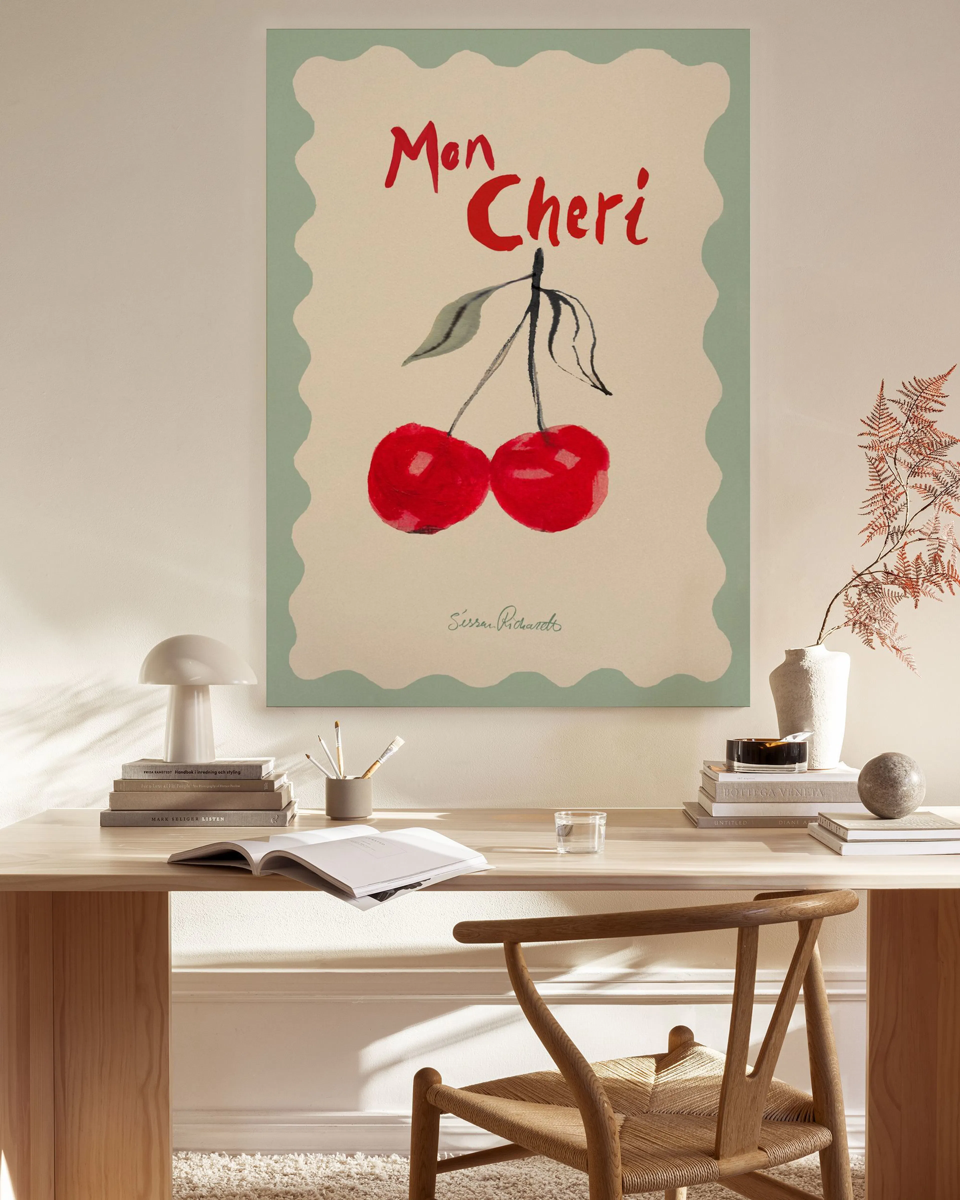 A canvas print of red cherries and Mon Cheri text on a light background with a green wavy border, hanging in a modern living roo