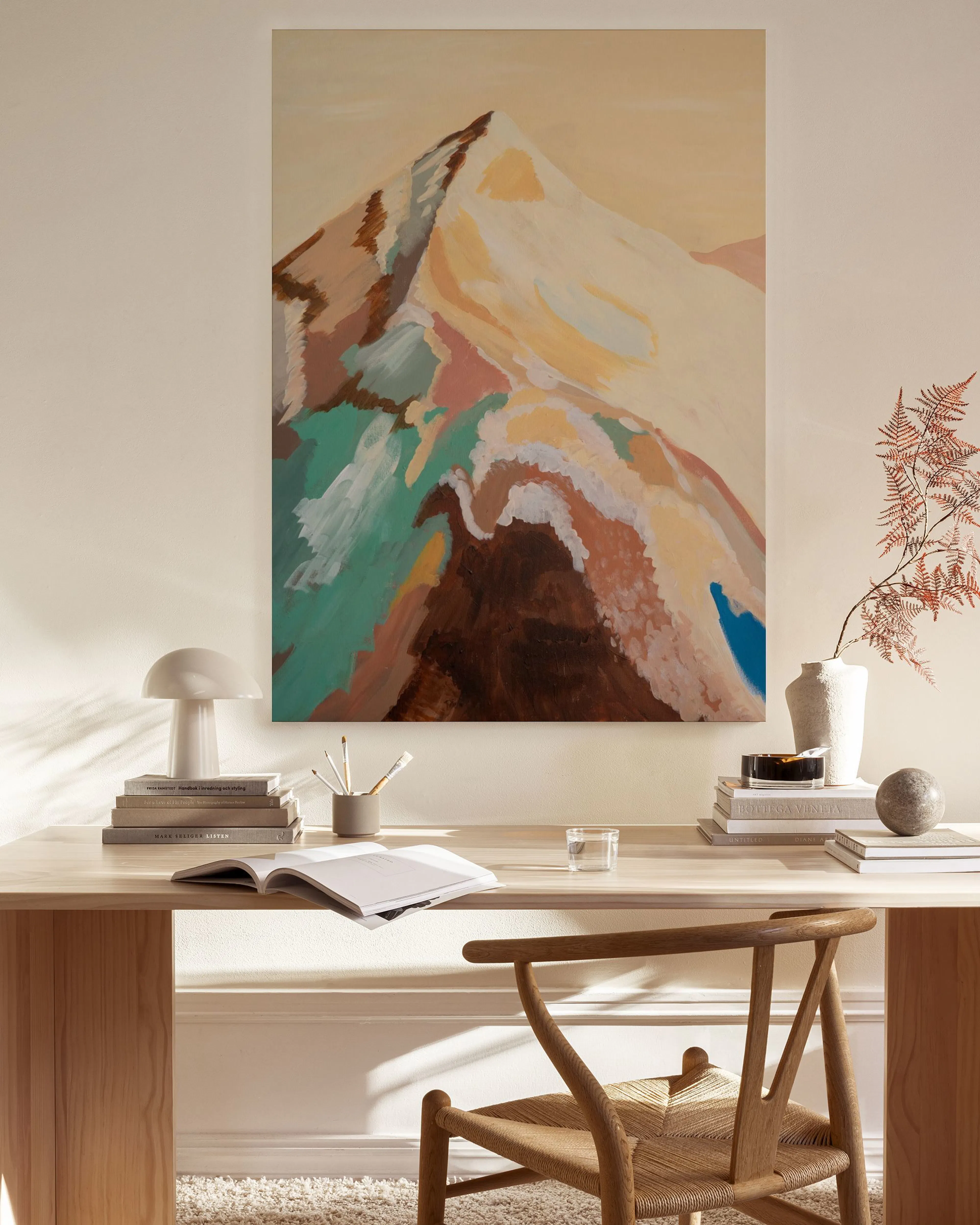 Lienzo abstracto de montaña en tonos tierra y azules, colgado sobre un mueble bajo, con una lámpara y un sillón de madera en un 