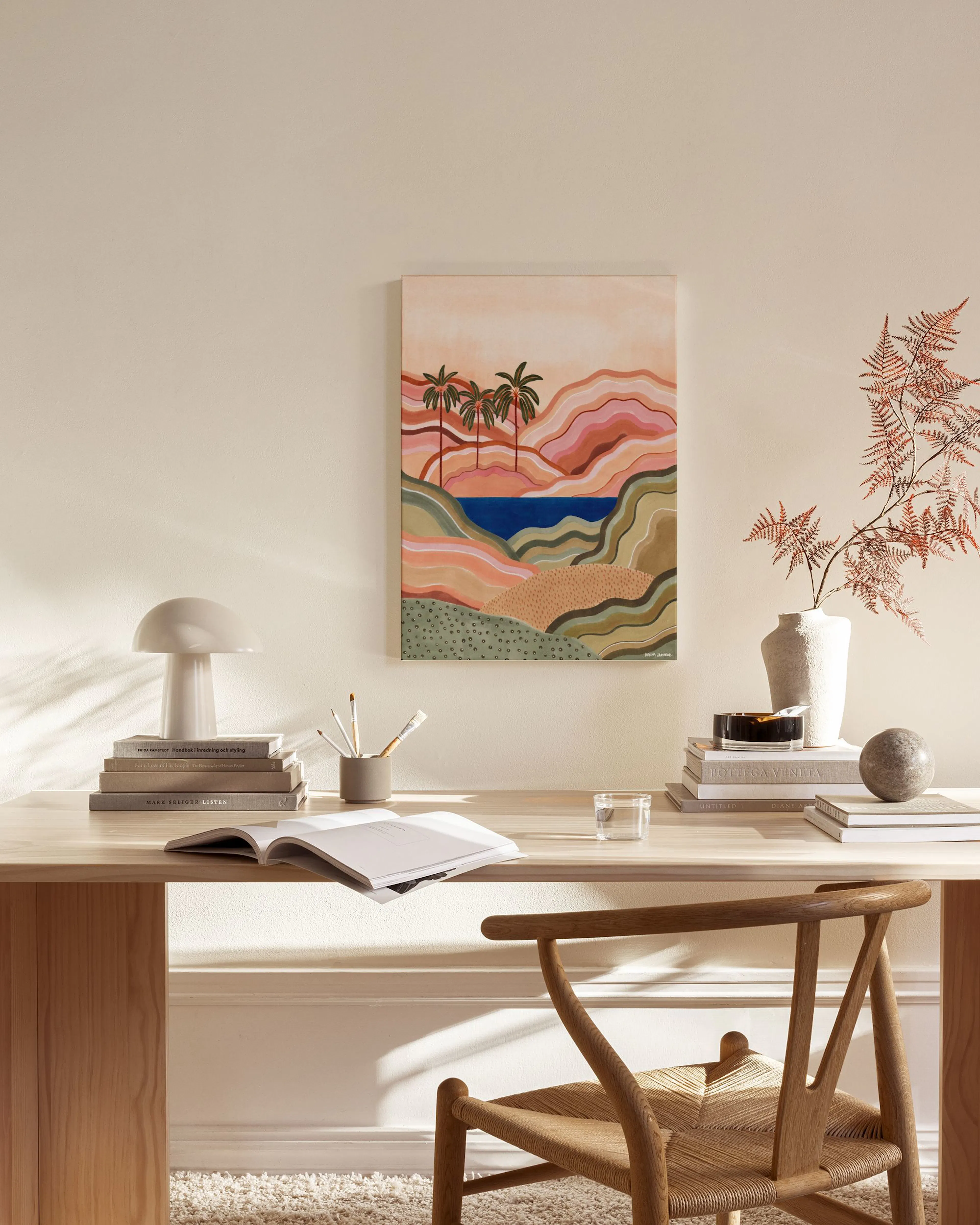 Abstract landscape canvas print with palm trees, rolling hills in pinks and greens, and a blue lake, hanging in a bright living 