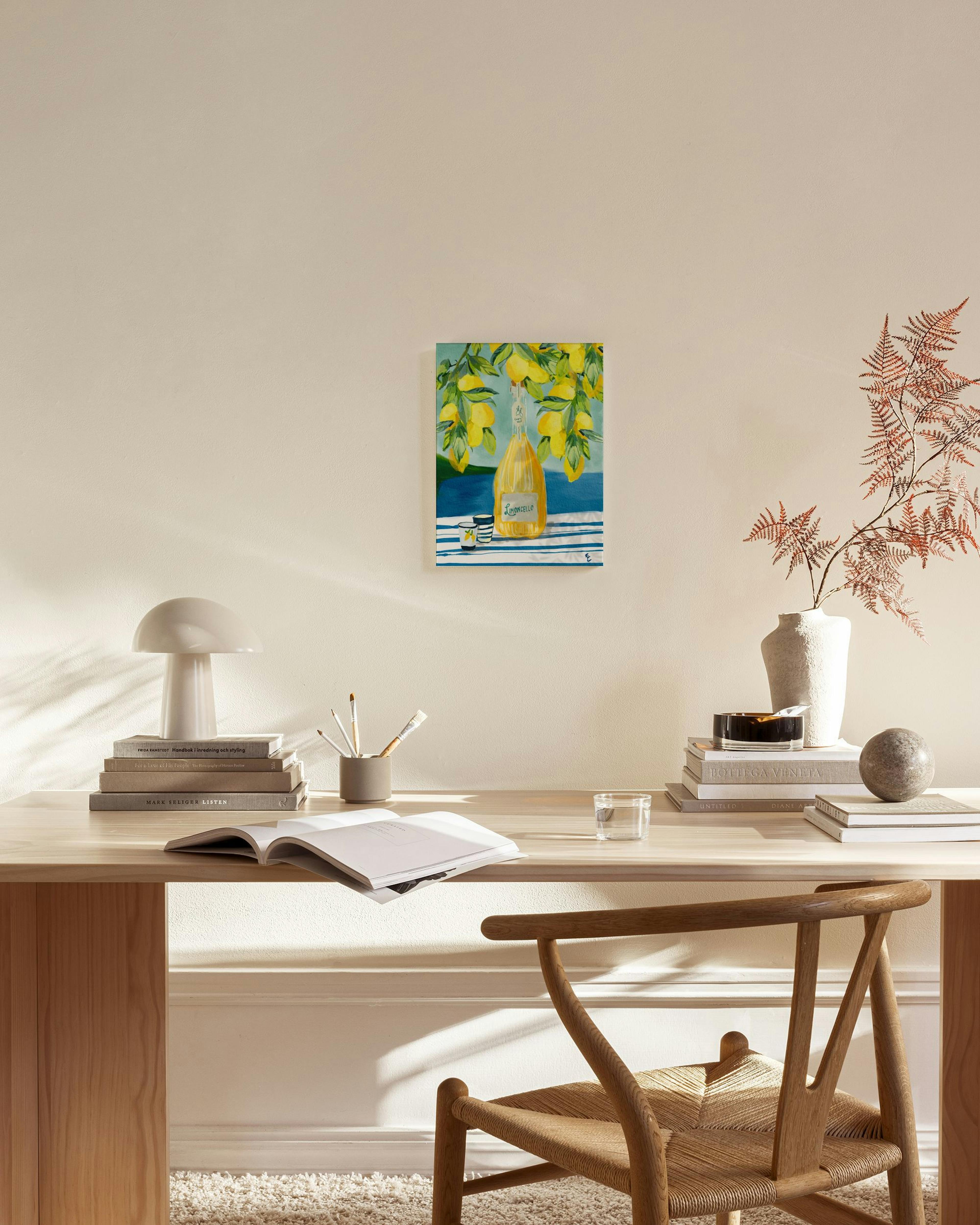 Καμβάς με μπουκάλι λιμοντσέλο, ποτήρια και κλαδιά λεμονιάς, κρεμασμένος σε μπεζ τοίχο πάνω από ξύλινο έπιπλο σε σαλόνι.