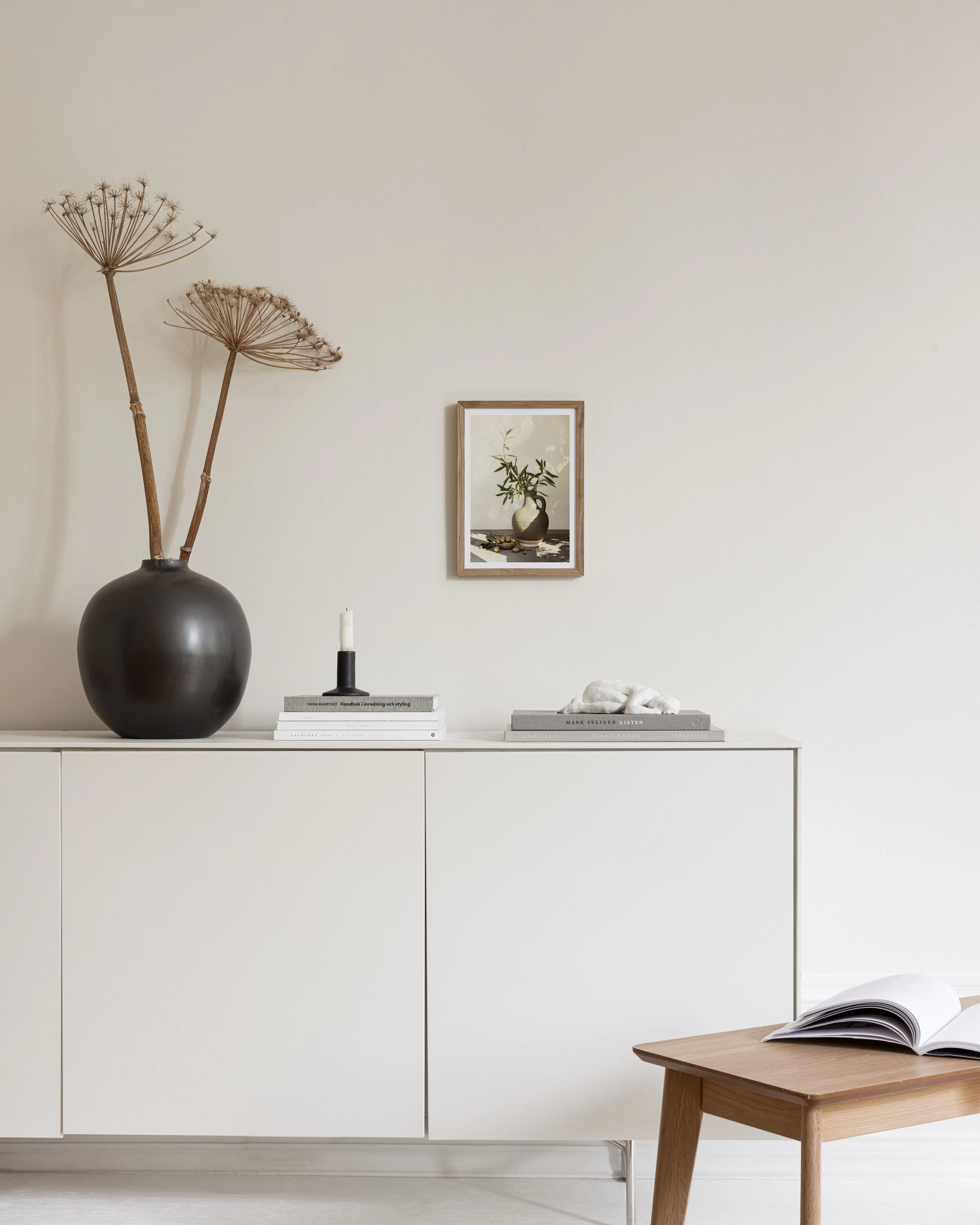 白い壁に飾られた、オリーブの枝が生けられた壺と実のポスター。下に白いキャビネットと黒い花瓶がある。