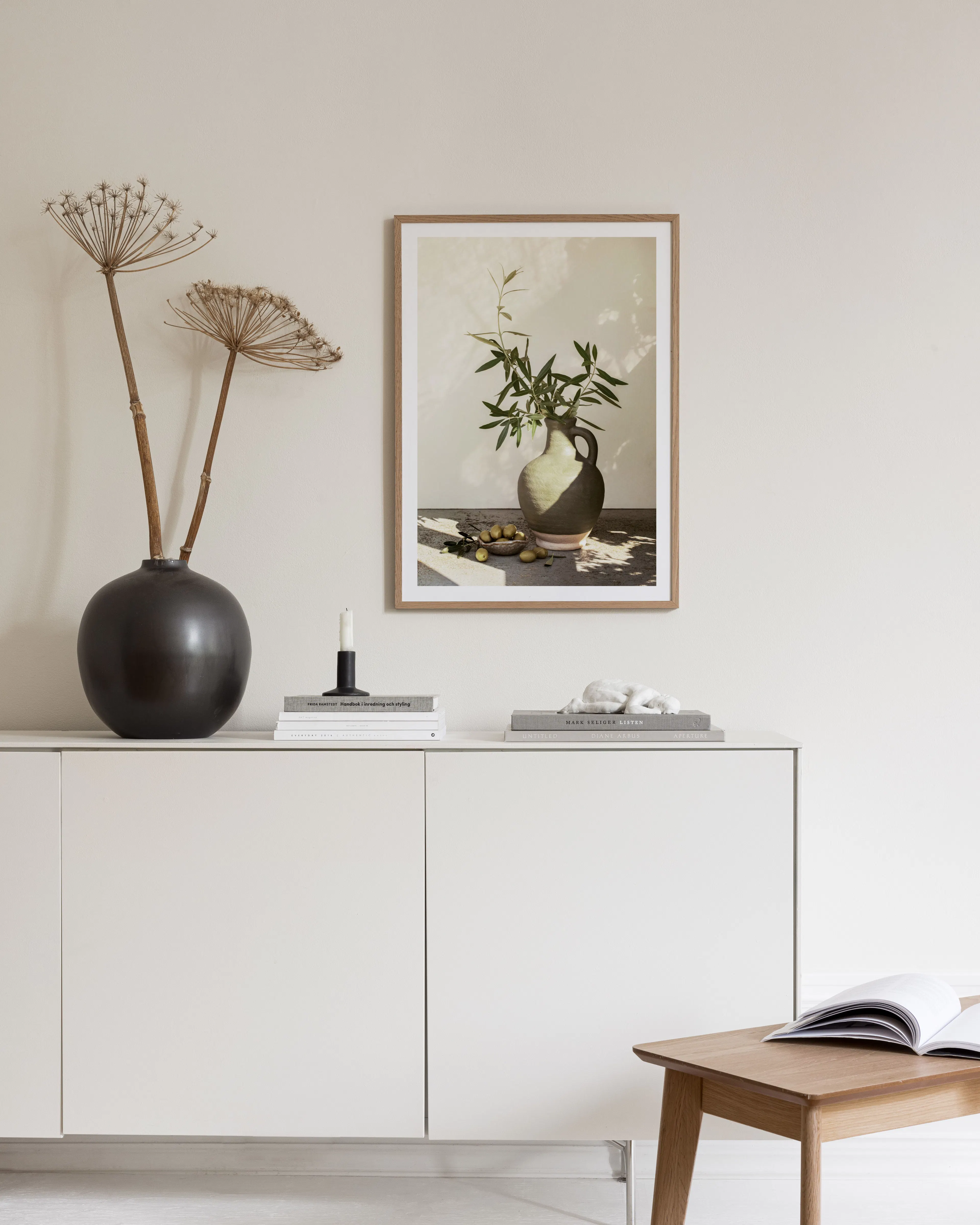 Poster van een vaas met olijftakken en olijven, hangend boven een wit dressoir met decoratie.