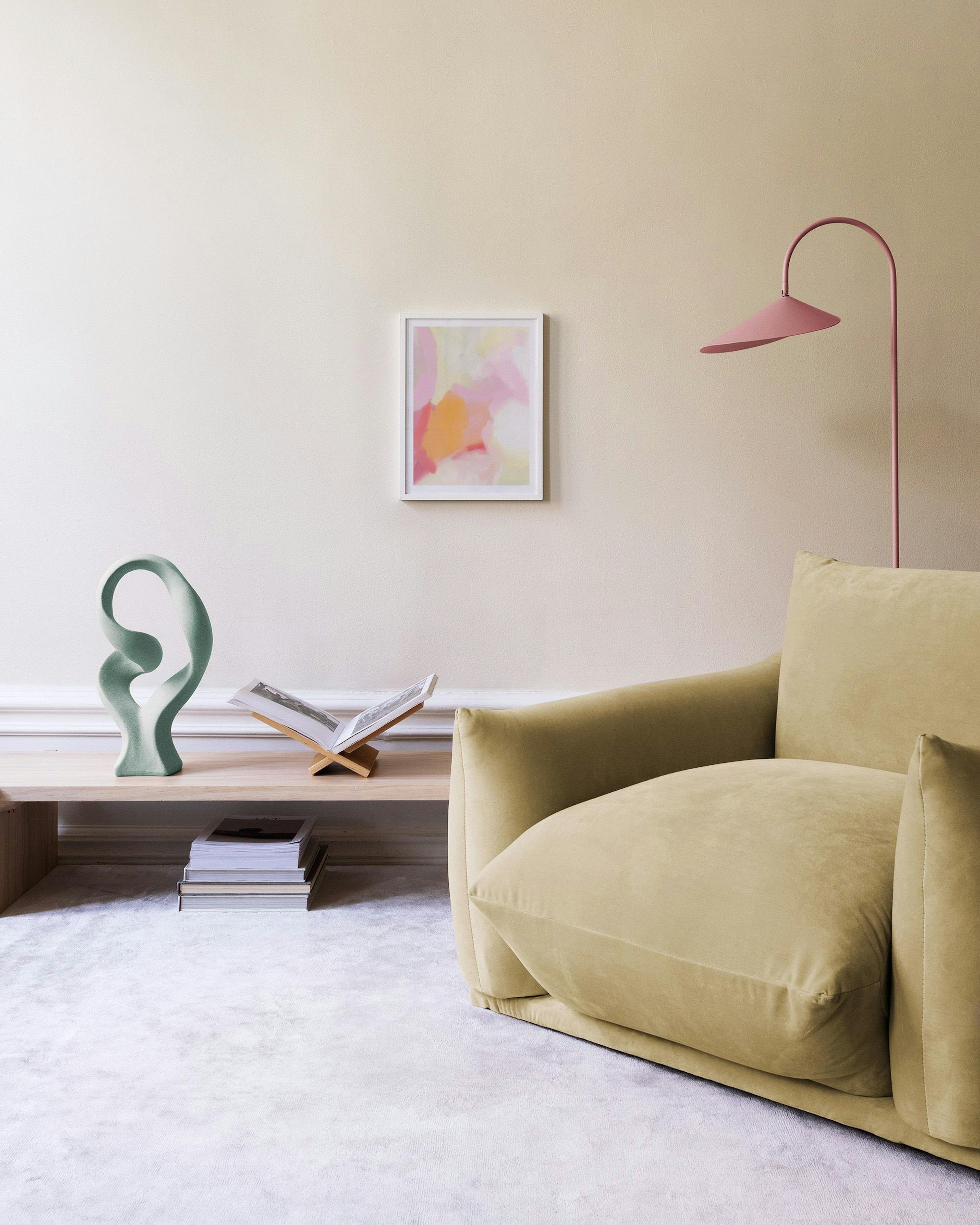 Abstrakt poster med rosa, orange och gula penseldrag på en beige vägg ovanför ett träskrivbord med bok och blomma.