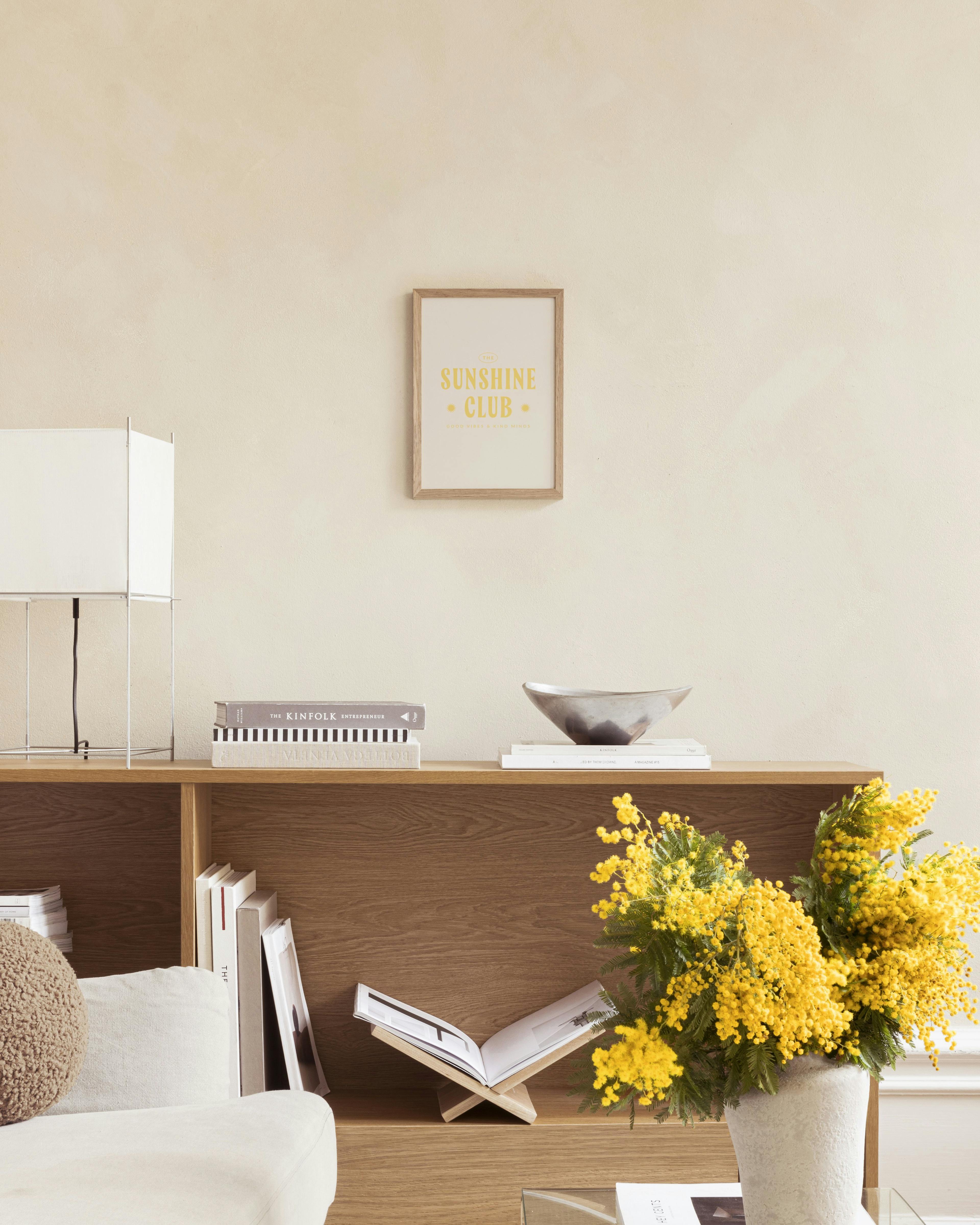 A print with THE SUNSHINE CLUB in yellow text on a beige background, hanging above a wooden shelf in a bright room.