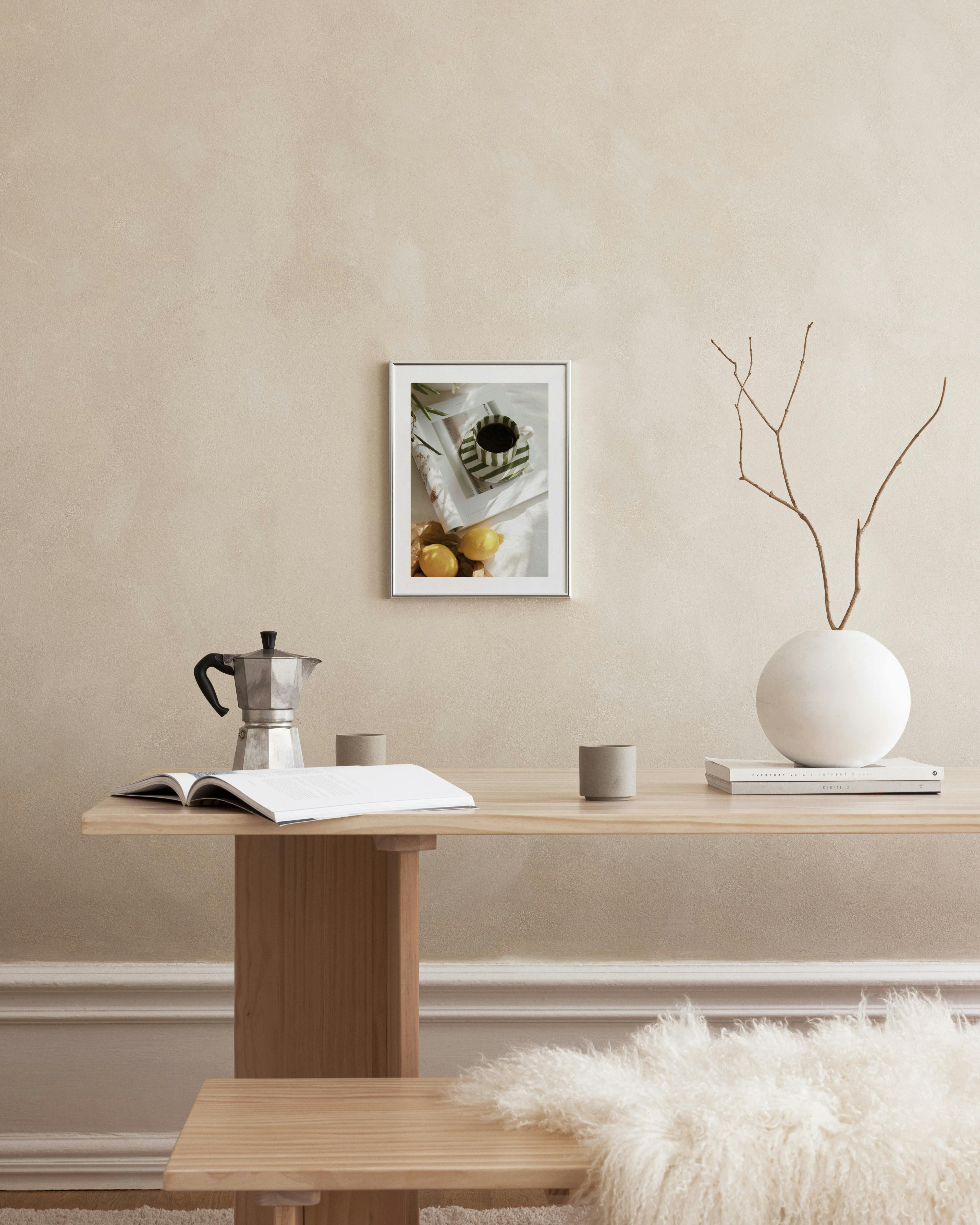 Πόστερ με φλιτζάνι καφέ με πράσινες ρίγες και λεμόνια, κρεμασμένο σε μπεζ τοίχο πάνω από ξύλινο τραπέζι.